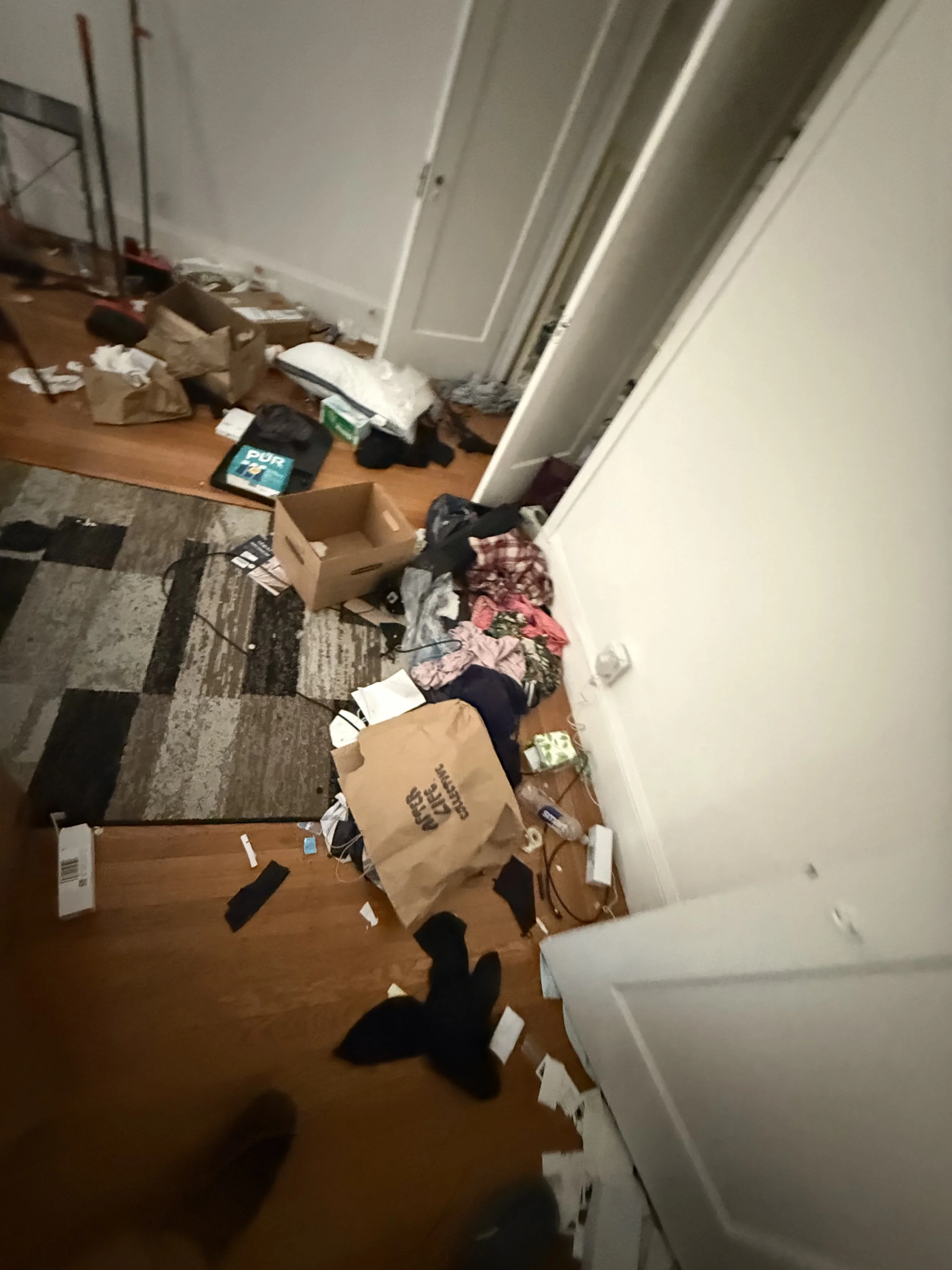 A cluttered room with scattered trash, cardboard boxes, papers, clothes, and miscellaneous items on a wood floor and a patterned rug, with a closed door and white walls.