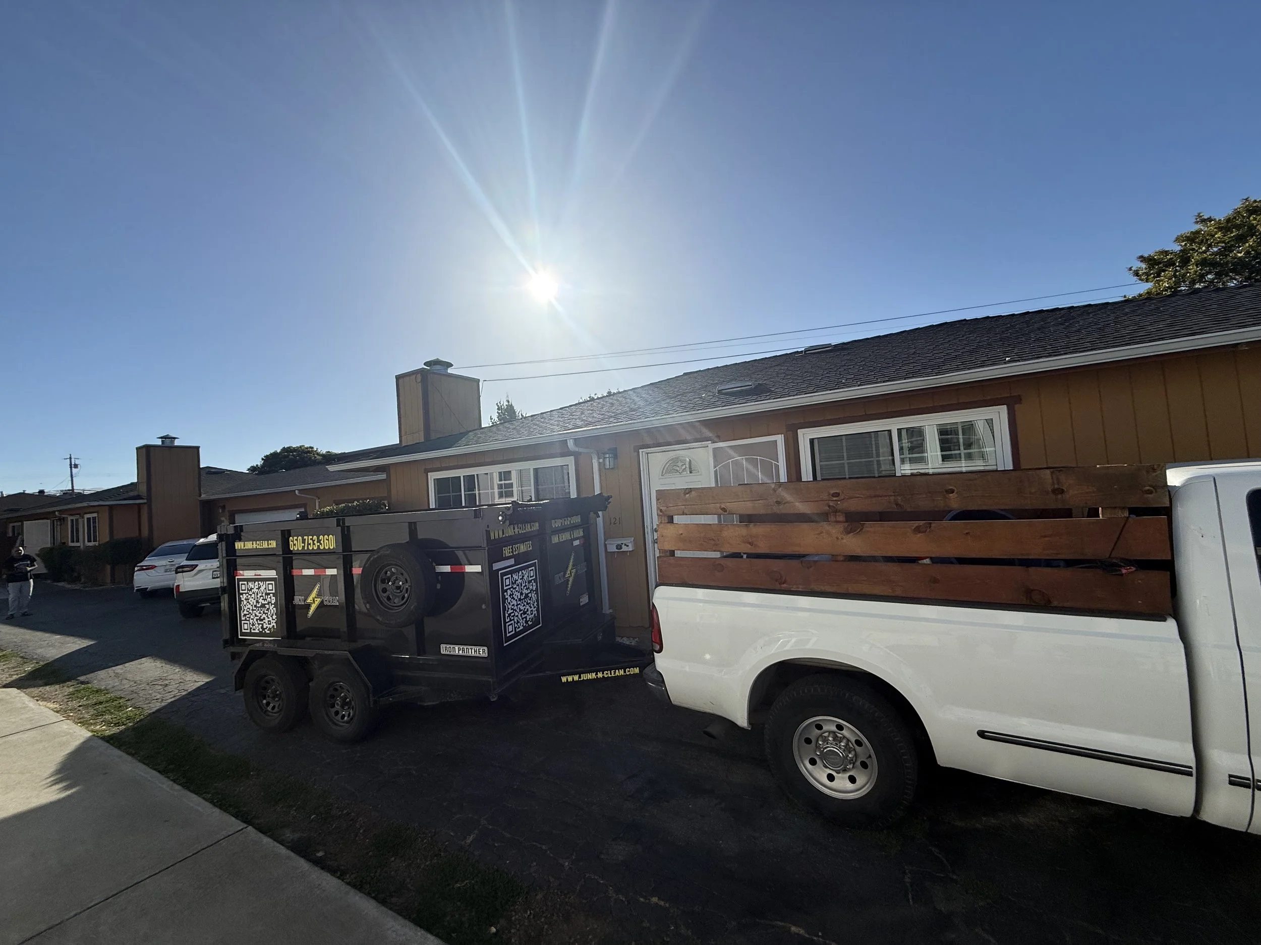 A sunny residential street with single-story houses, parked cars, and a black trailer with QR codes on it. The sky is clear and blue with the sun shining brightly.