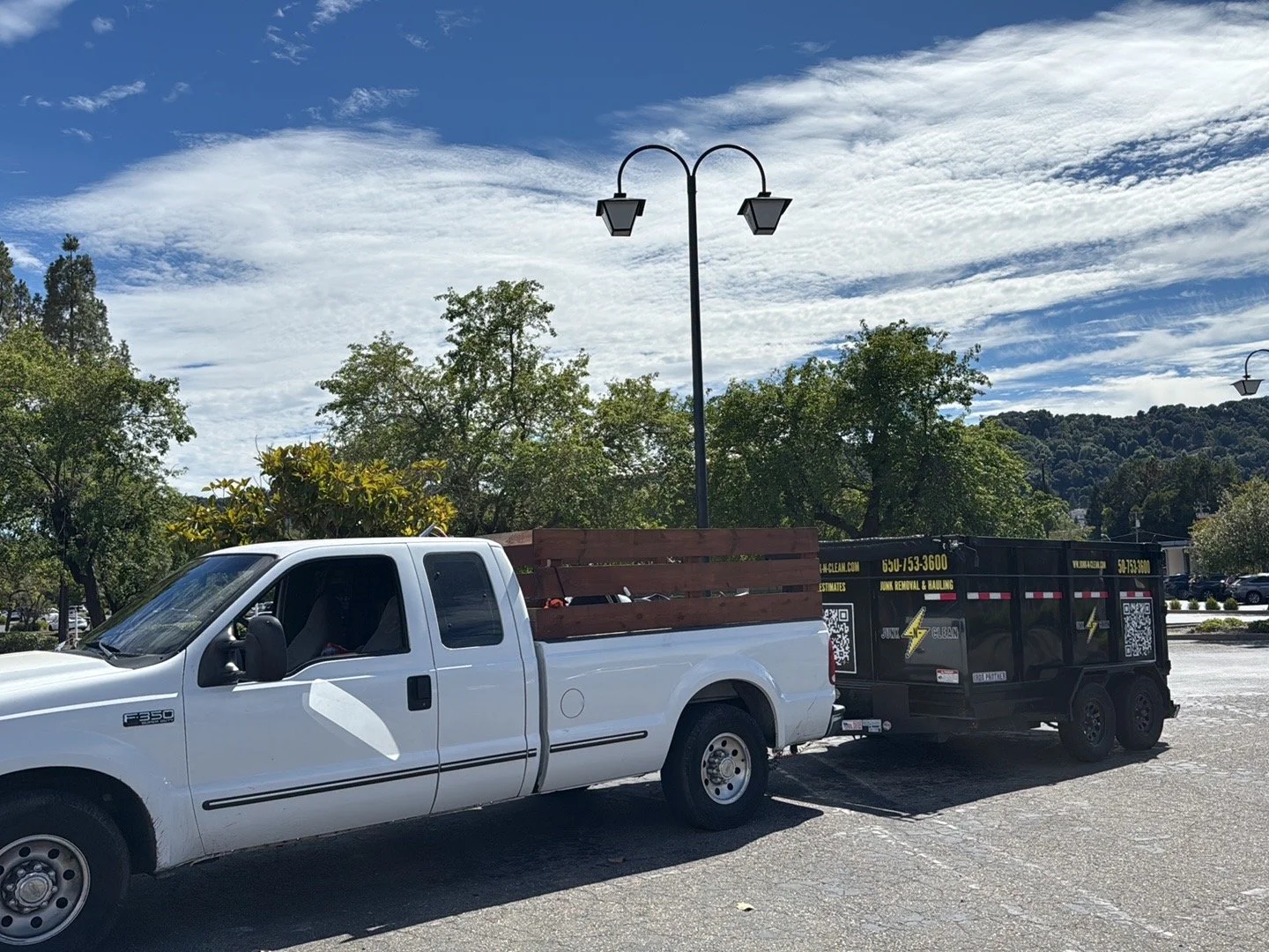 White Ford pickup truck towing a black dump trailer in a parking lot with trees and mountains in the background under a partly cloudy sky.