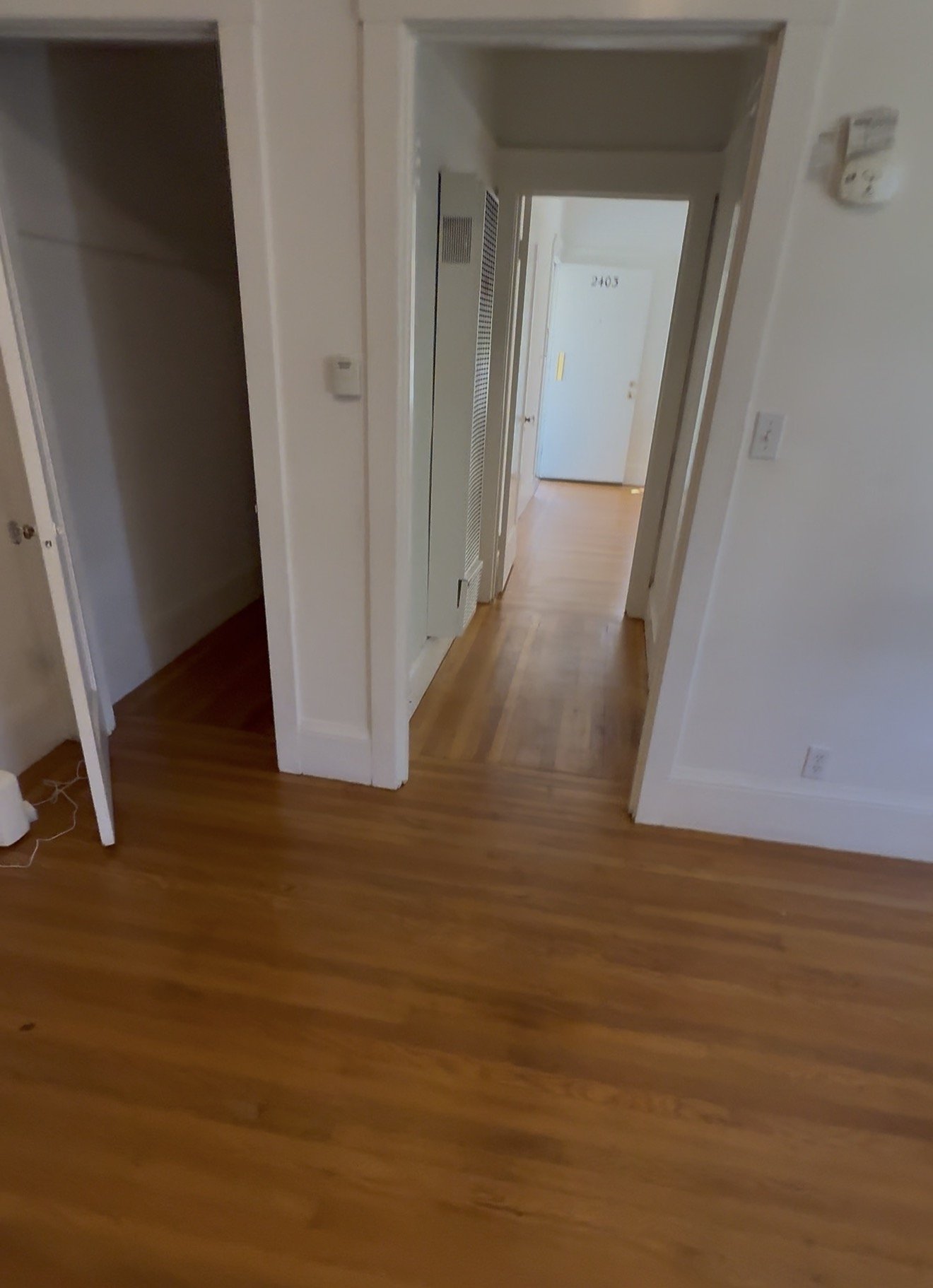 View of an interior hallway in an apartment with wooden flooring, white walls, a doorway on the left, a thermostat on the wall on the right, and doors leading to rooms with visible door numbers.