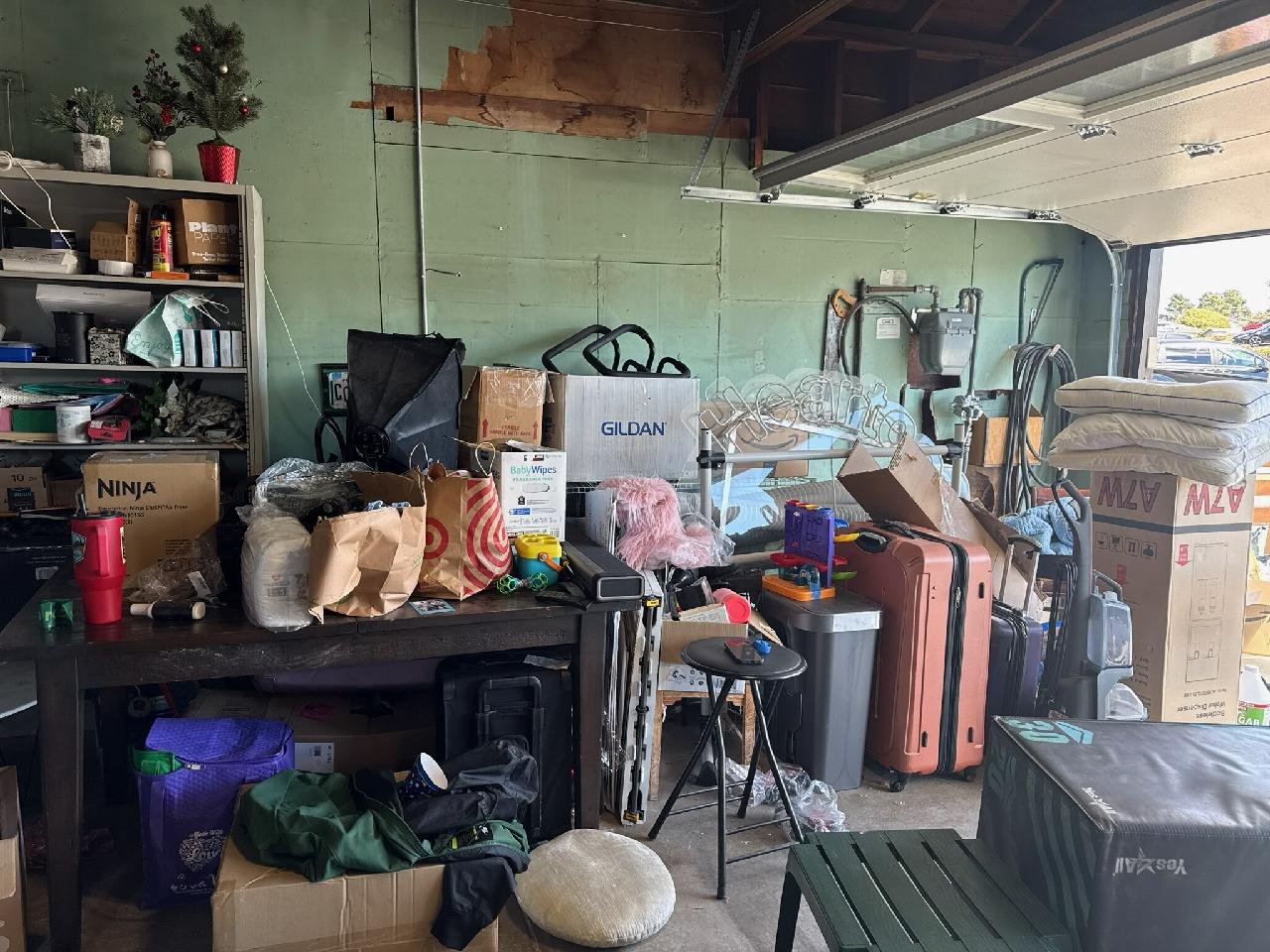 A cluttered garage with various boxes, bags, and household items. There are suitcases, a small table with toys, a shelf with storage bins, and a partially open garage door.