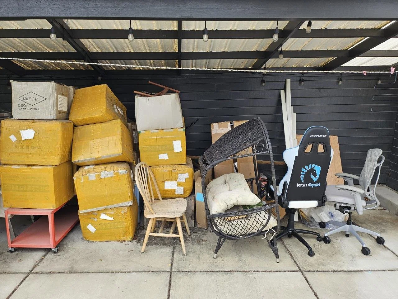 A storage area with stacked cardboard boxes, chairs, and office equipment under a metal roof with string lights.