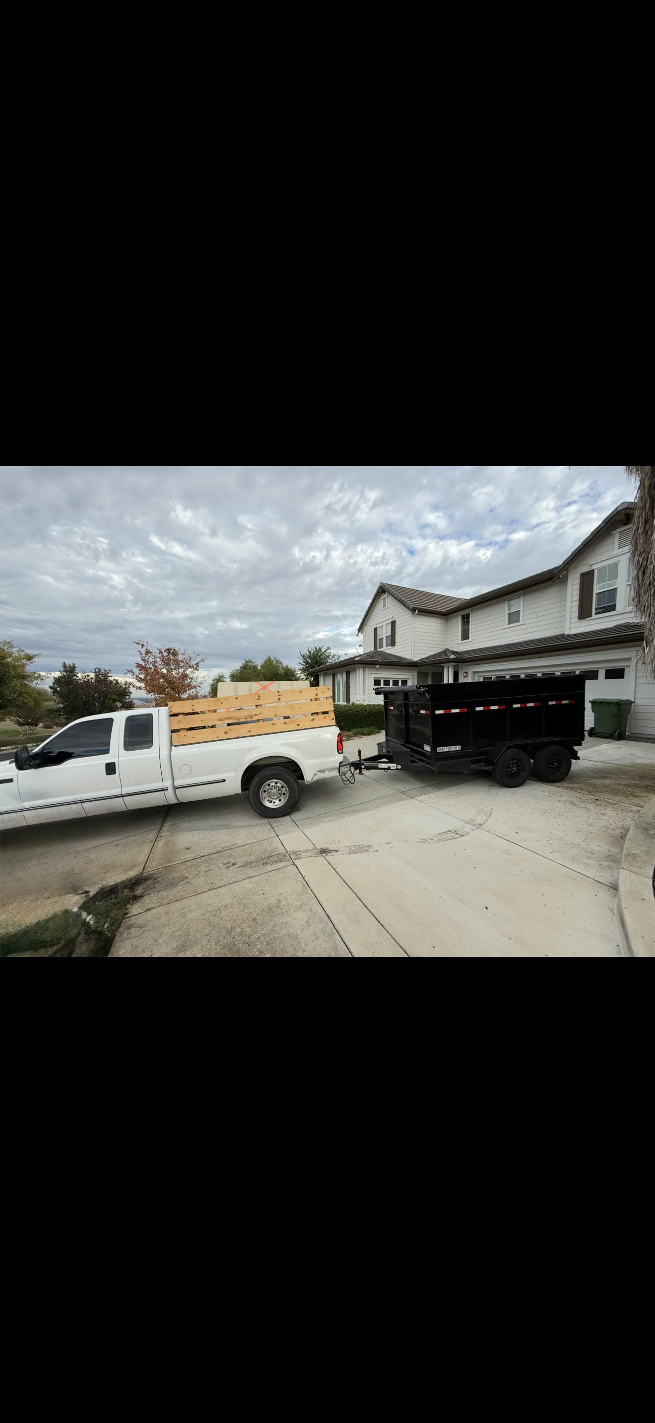 A white pickup truck with wooden planks in the bed, attached to a black utility trailer parked in front of a suburban house with a driveway, trees, and cloudy sky.