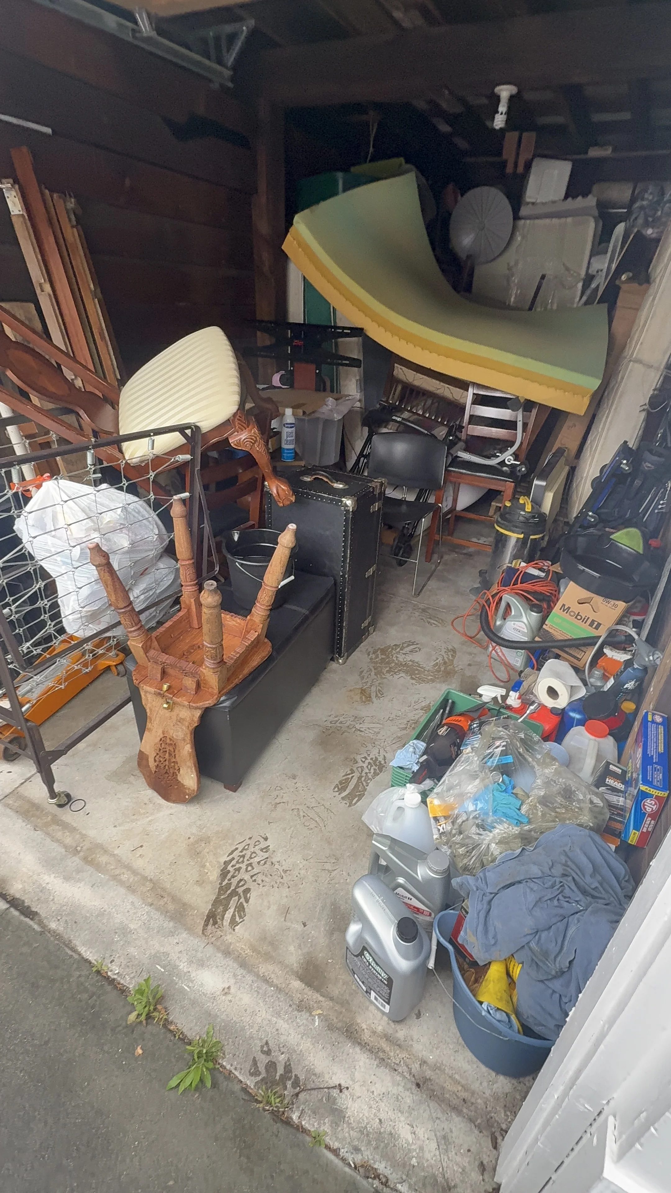 Garage filled with various household items and tools, including chairs, a small black suitcase, cleaning supplies, motor oil, and plastic containers. There are foot prints on the concrete floor and a small green plant at the edge of the garage.