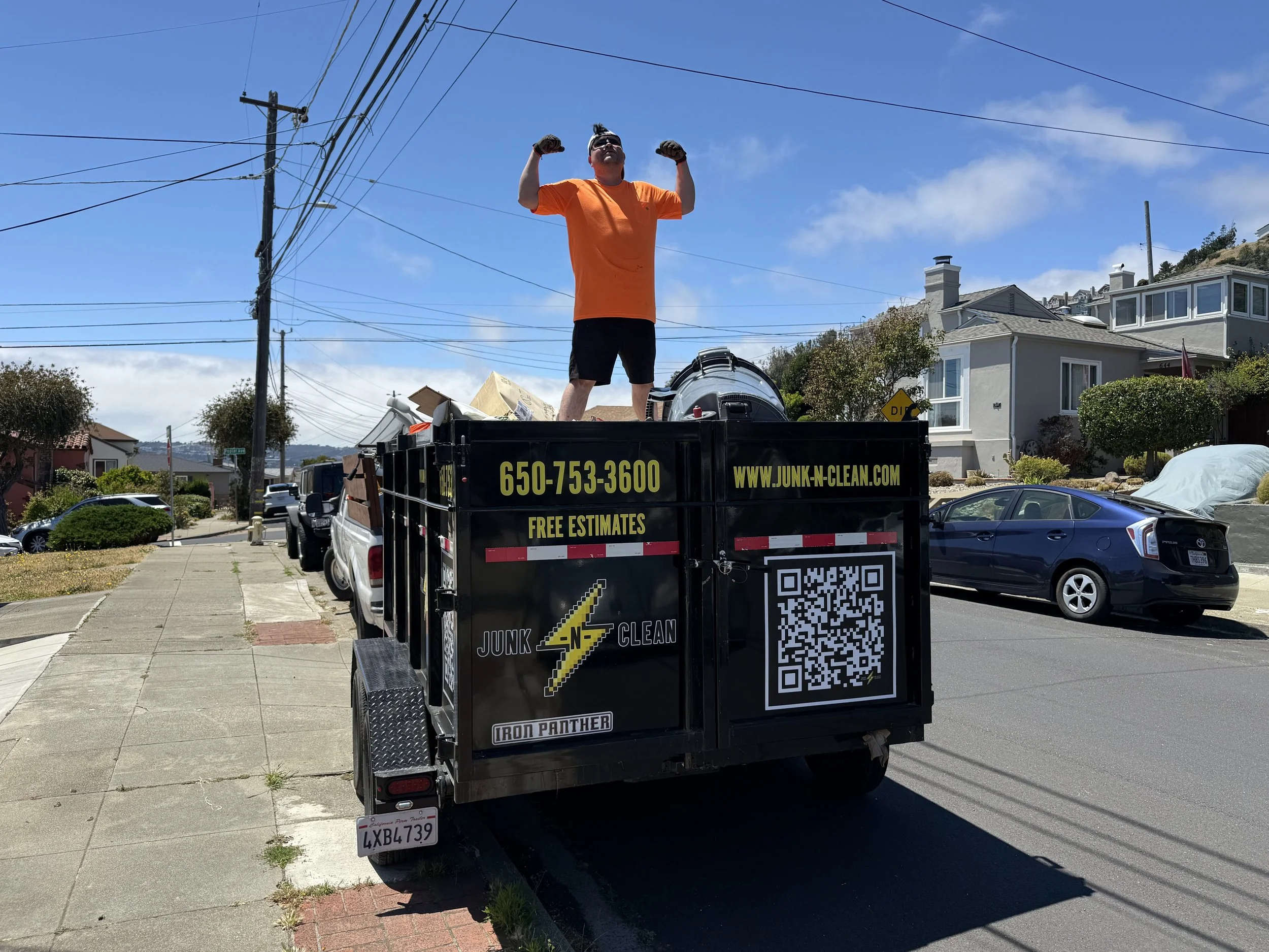 A man standing in a truck bed with his arms flexed, wearing an orange shirt and sunglasses, on a street with houses and power lines under a blue sky.