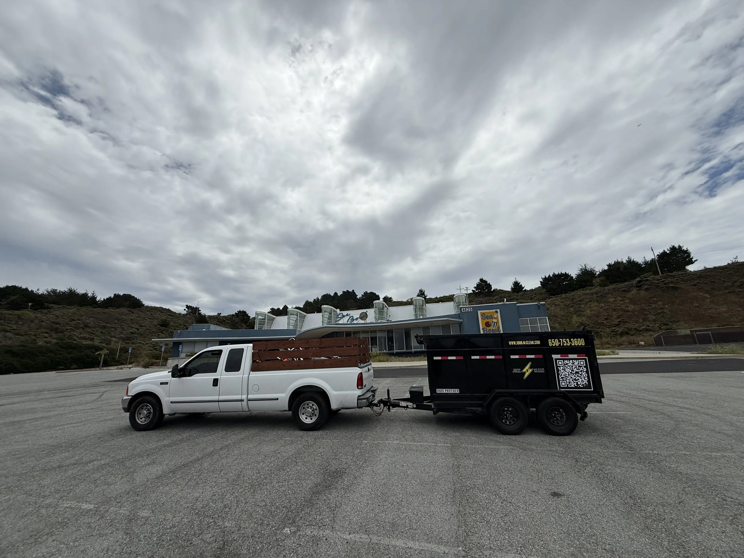 A white pickup truck is towing a black trailer with a QR code and company information, parked in an empty lot under a cloudy sky, with a building labeled 'Sea Bowl' in the background.