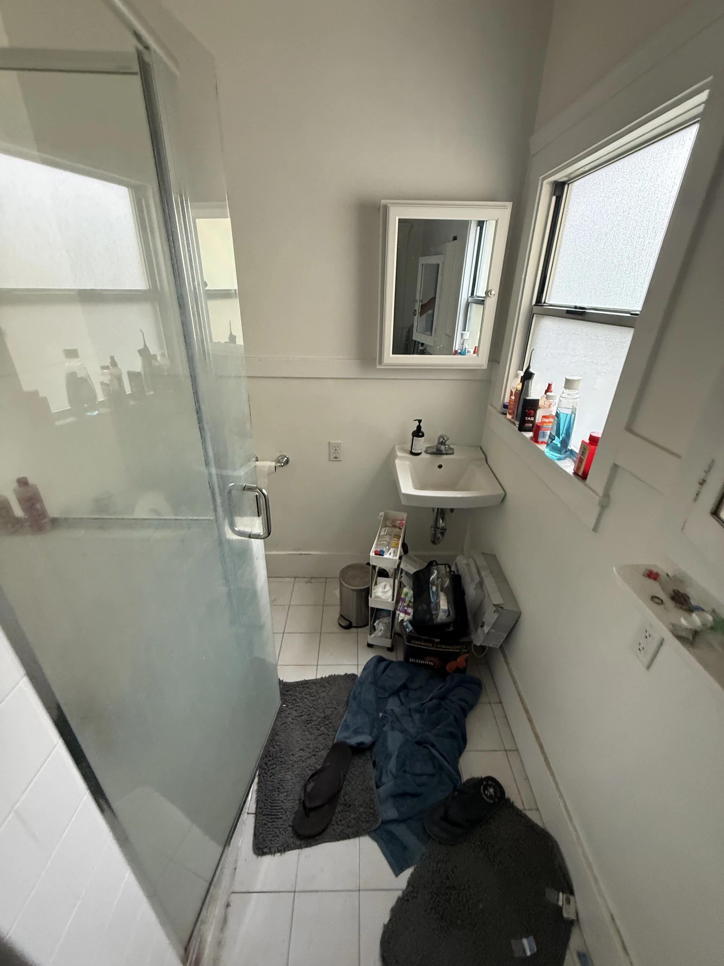 View of a cluttered bathroom with a person lying on the floor, various toiletries on the window sill, a mirror, and a shower stall with glass door.