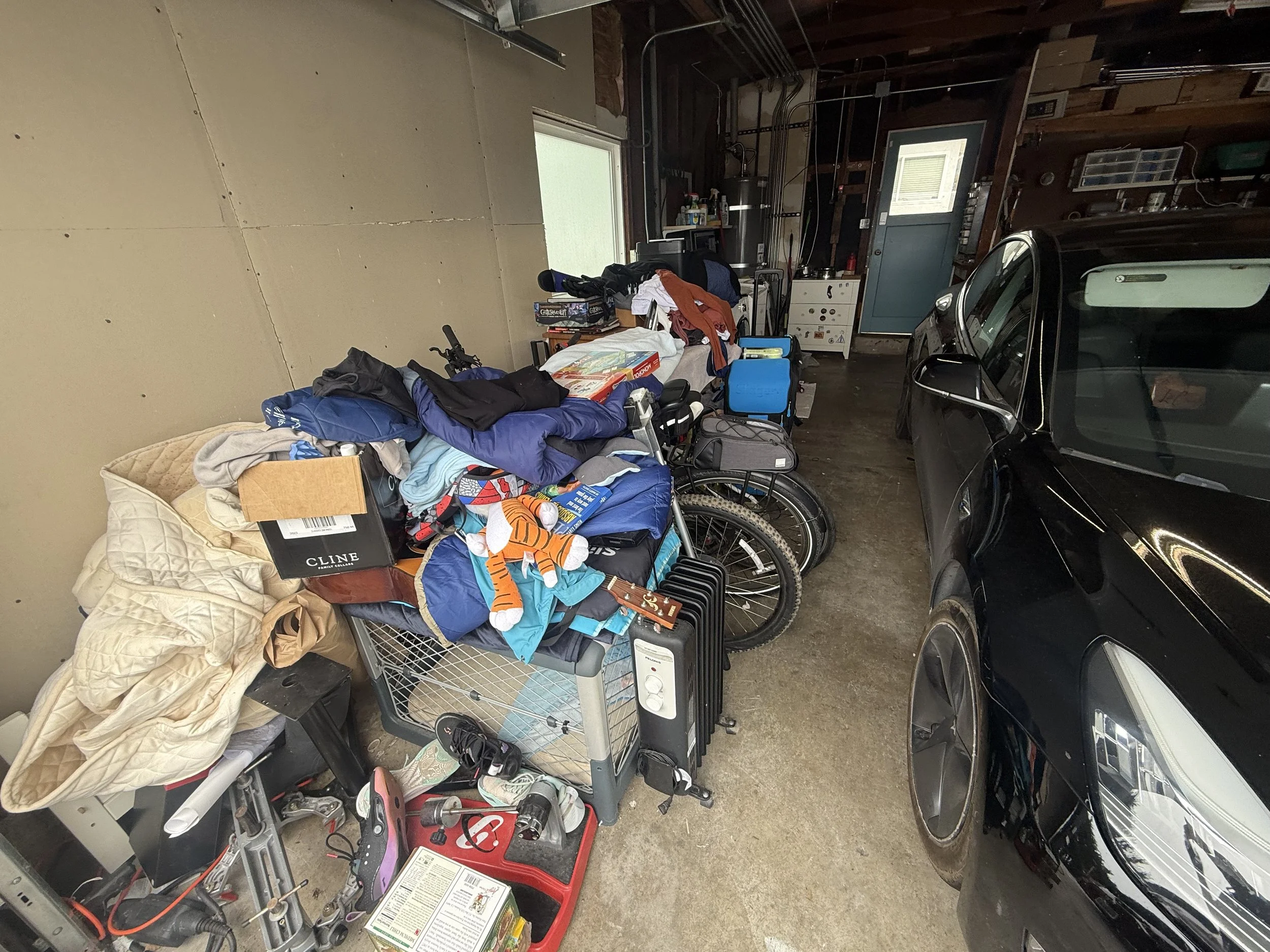 A cluttered garage with a table covered in clothes, boxes, and household items. A bicycle and a car are parked inside, with various tools and supplies on shelves and countertops.