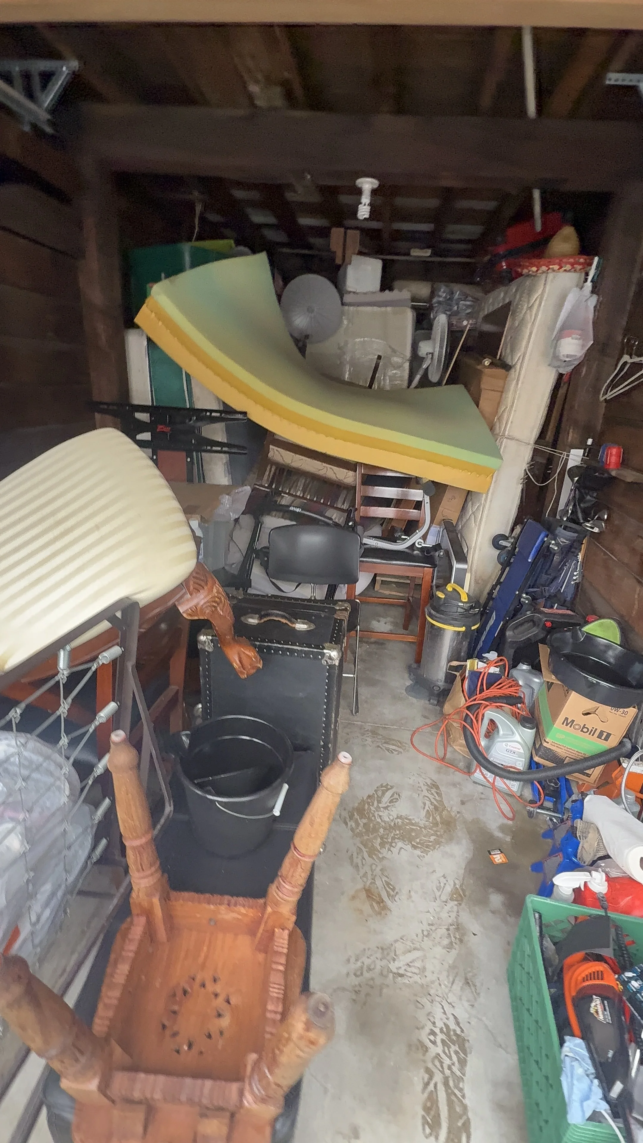 A cluttered garage with various household items, furniture, and tools stacked and scattered throughout, including chairs, a foam mattress, vacuum cleaner, and power tools.