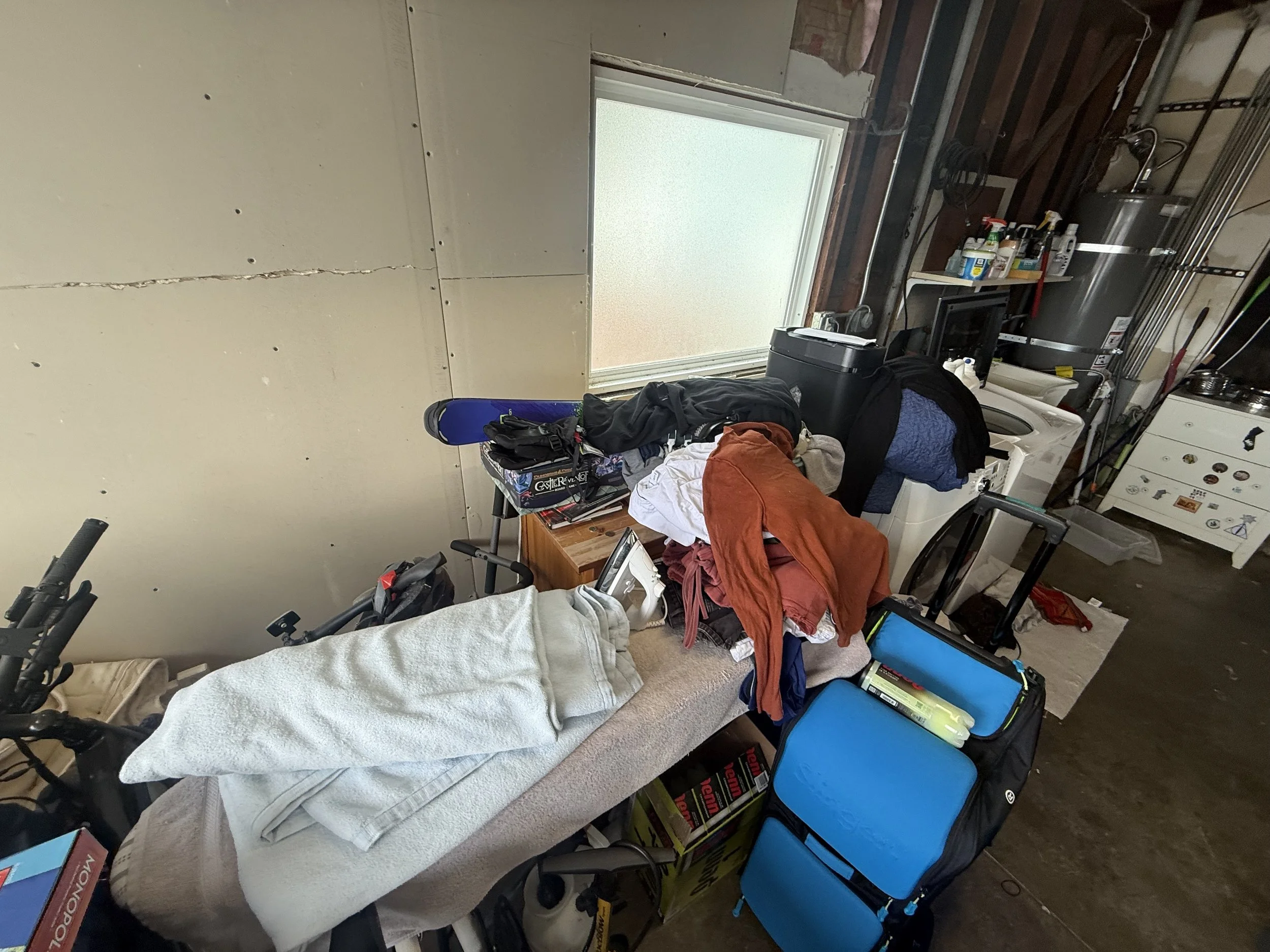 A cluttered garage with a bed covered in clothes and various items, including a blue foam roller, a black suitcase, and a chair, near a window with frosted glass. The garage has unfinished walls, pipes, shelves with cleaning supplies, and a washing m
