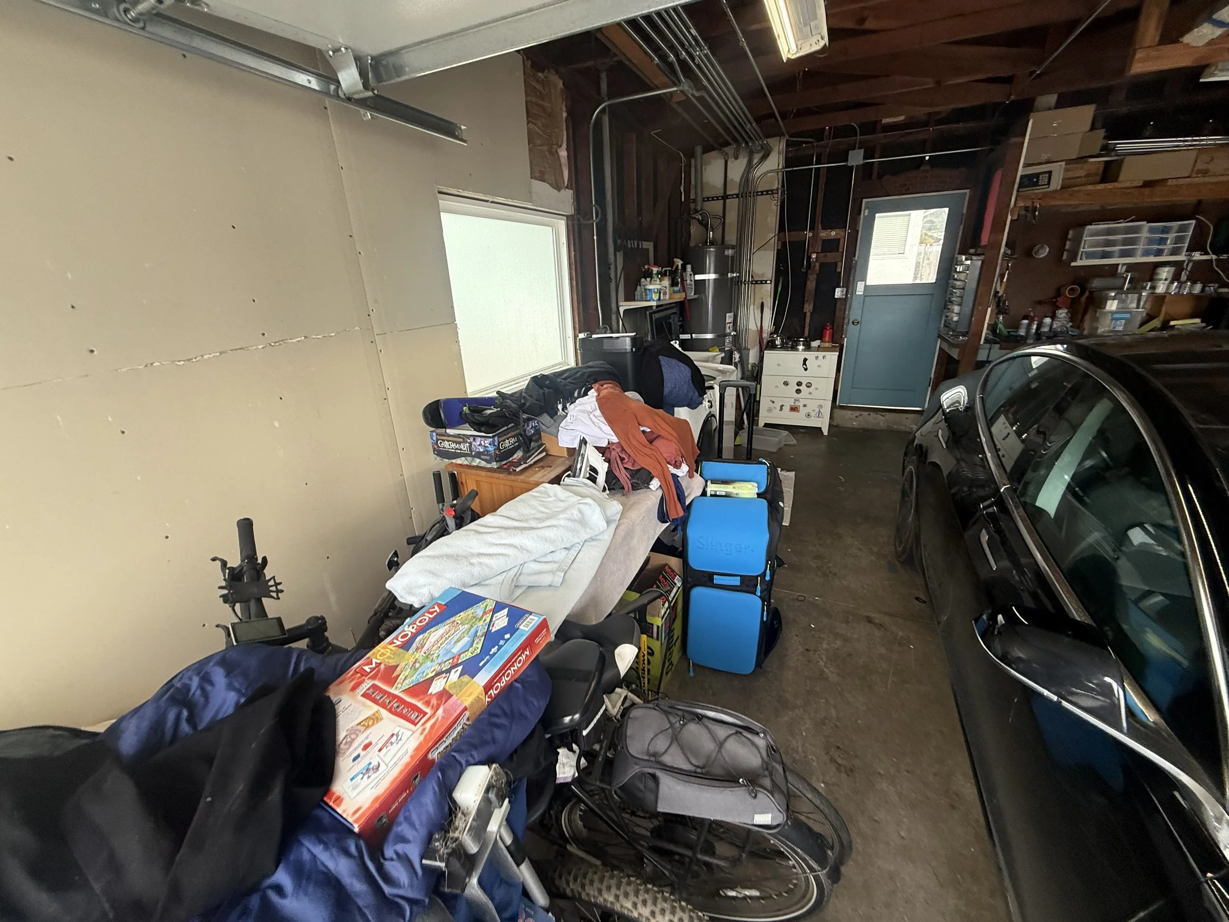 A cluttered garage with a black car parked inside, various items on a table, and a sofa with clothes and a board game box on top. There are tools, boxes, and household items along the walls and a small window providing natural light.