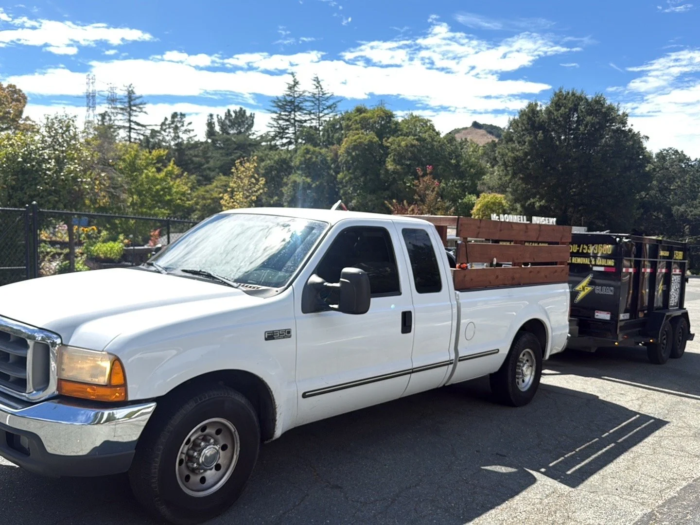 White Ford F-350 pickup truck towing a small black trailer with yellow and white text and a logo, parked on a sunny day with trees and a blue sky in the background.