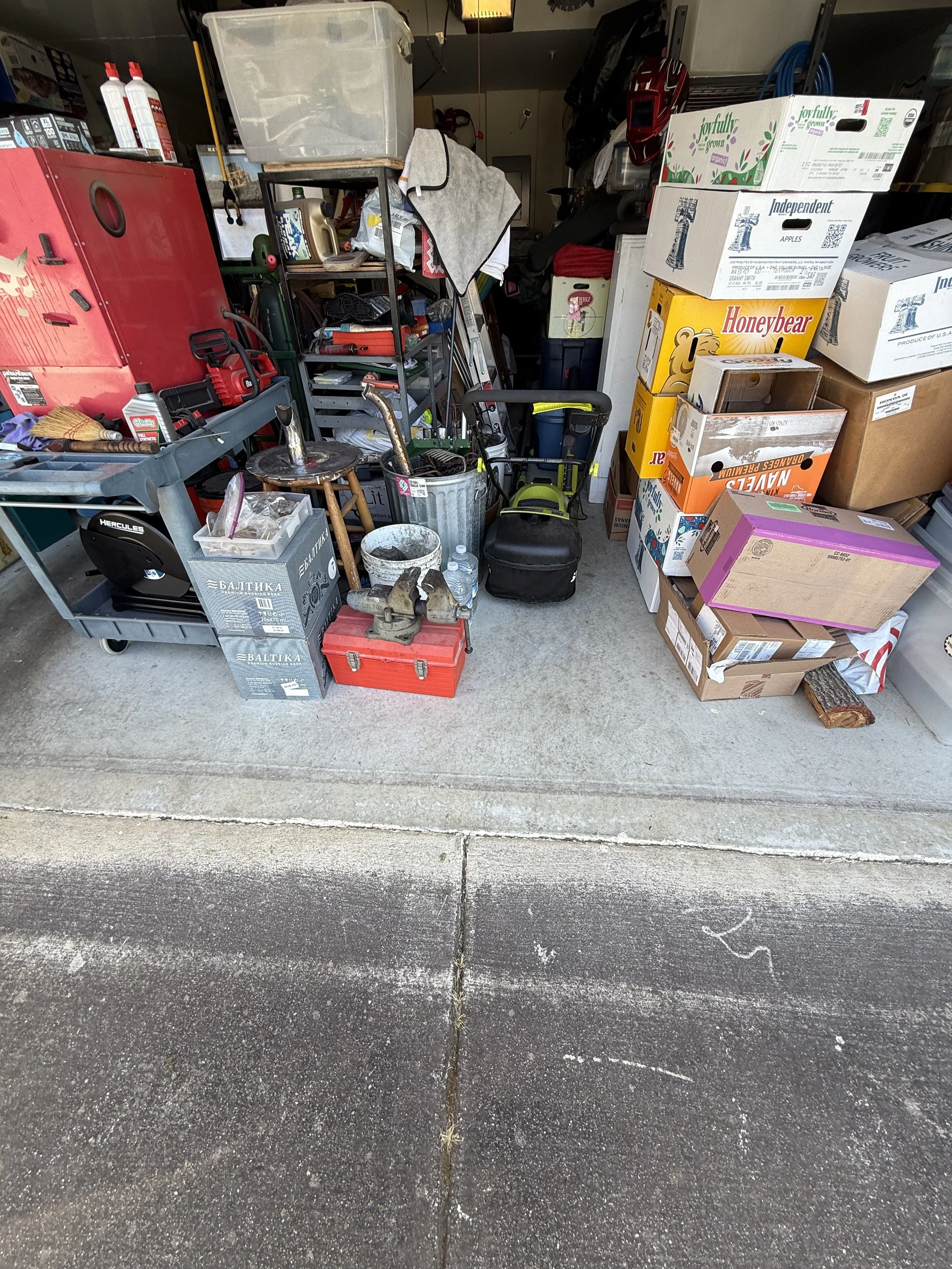 Garage with various stored items including boxes, tools, and household supplies. The foreground shows a concrete driveway with some chalk markings.