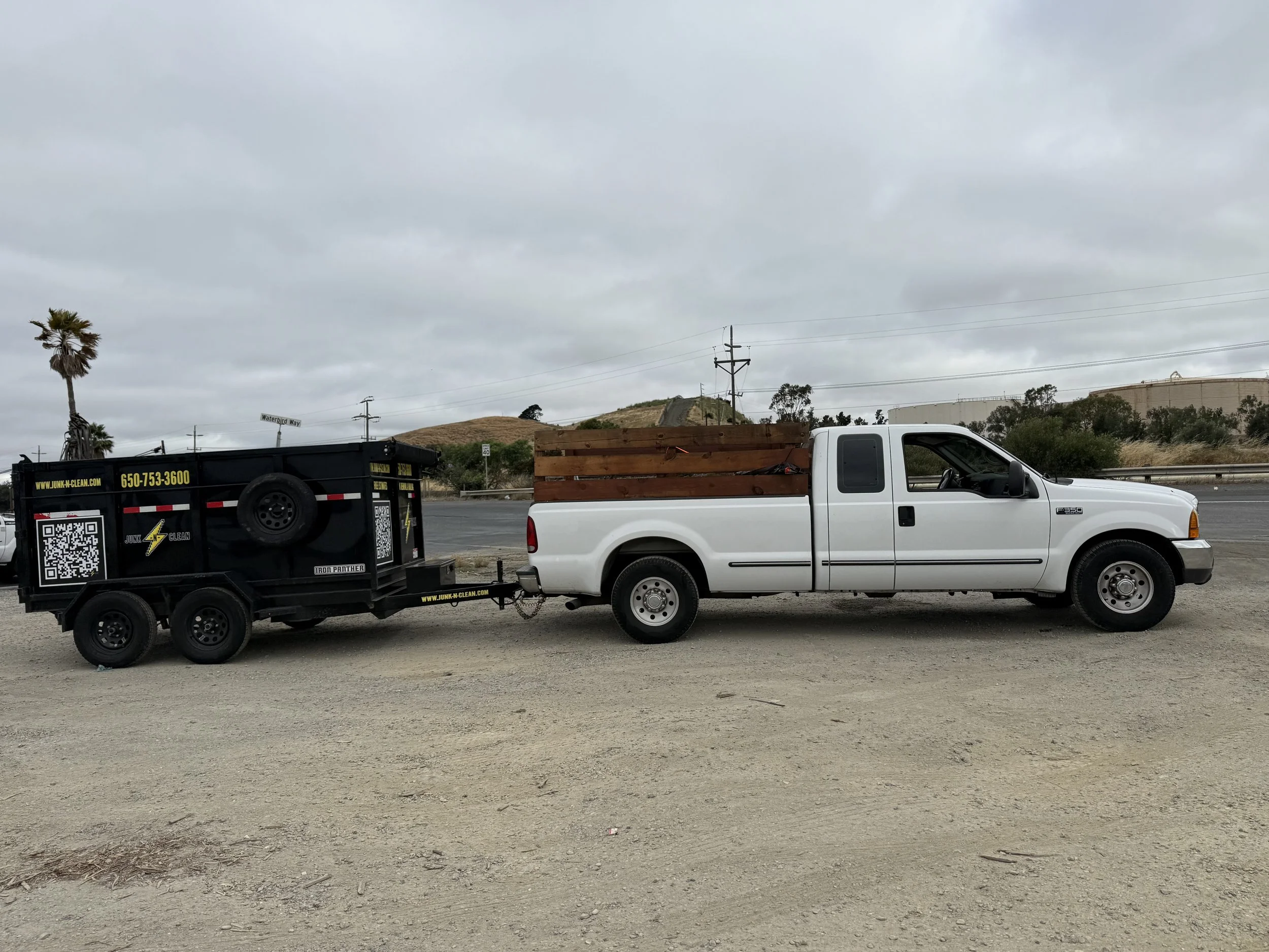 A white pickup truck attached to a black trailer with a website and phone number, parked on a dirt area near a road. In the background, there are hills, utility poles, and overcast sky.