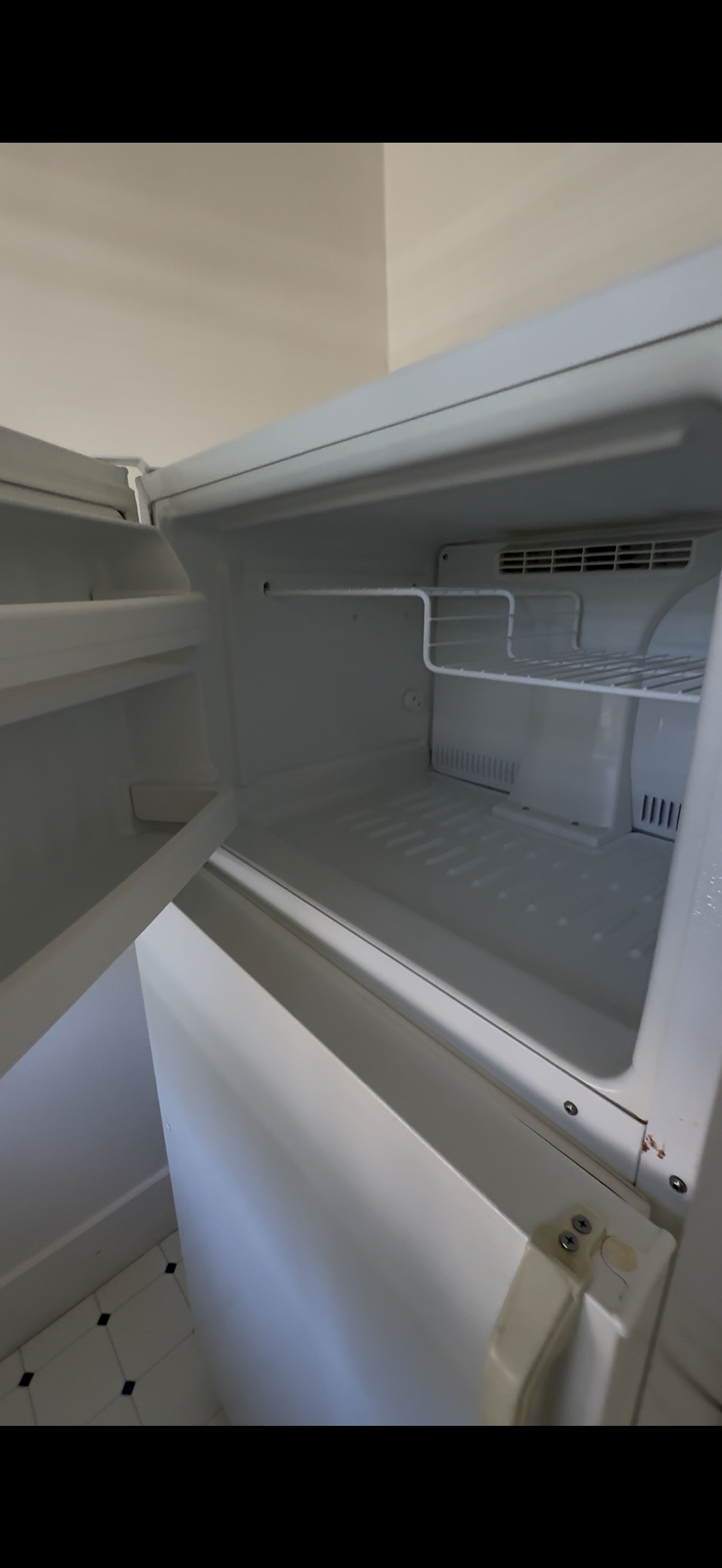 Empty white refrigerator with open door showing internal shelves and a wire rack, situated in a kitchen with black and white tiled flooring.