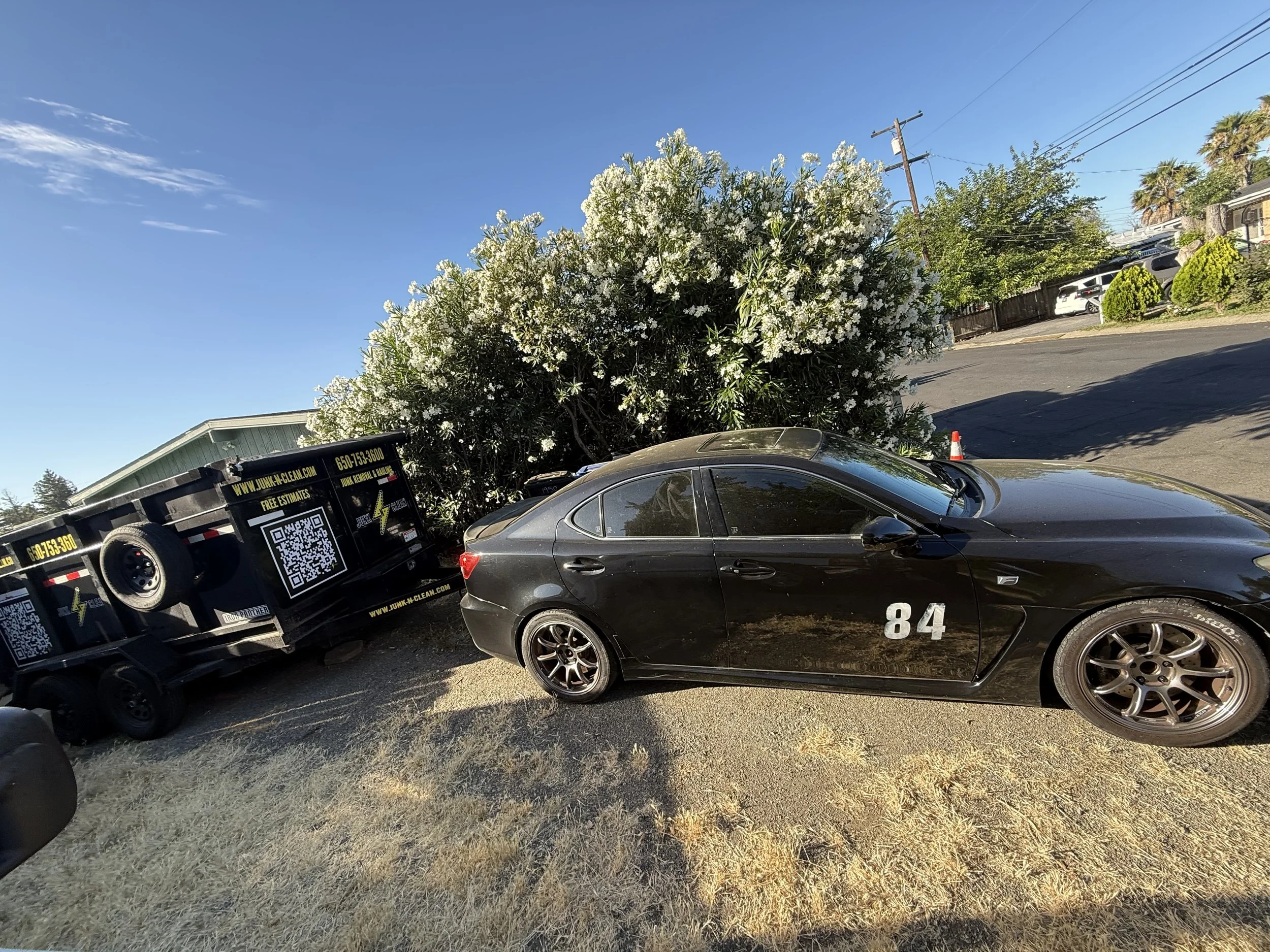 A black sports car with the number 84 on the side parked next to a tree with white flowers. A black utility truck with advertising and QR code on the side is parked behind the car. The background includes residential houses, palm trees, power lines, 