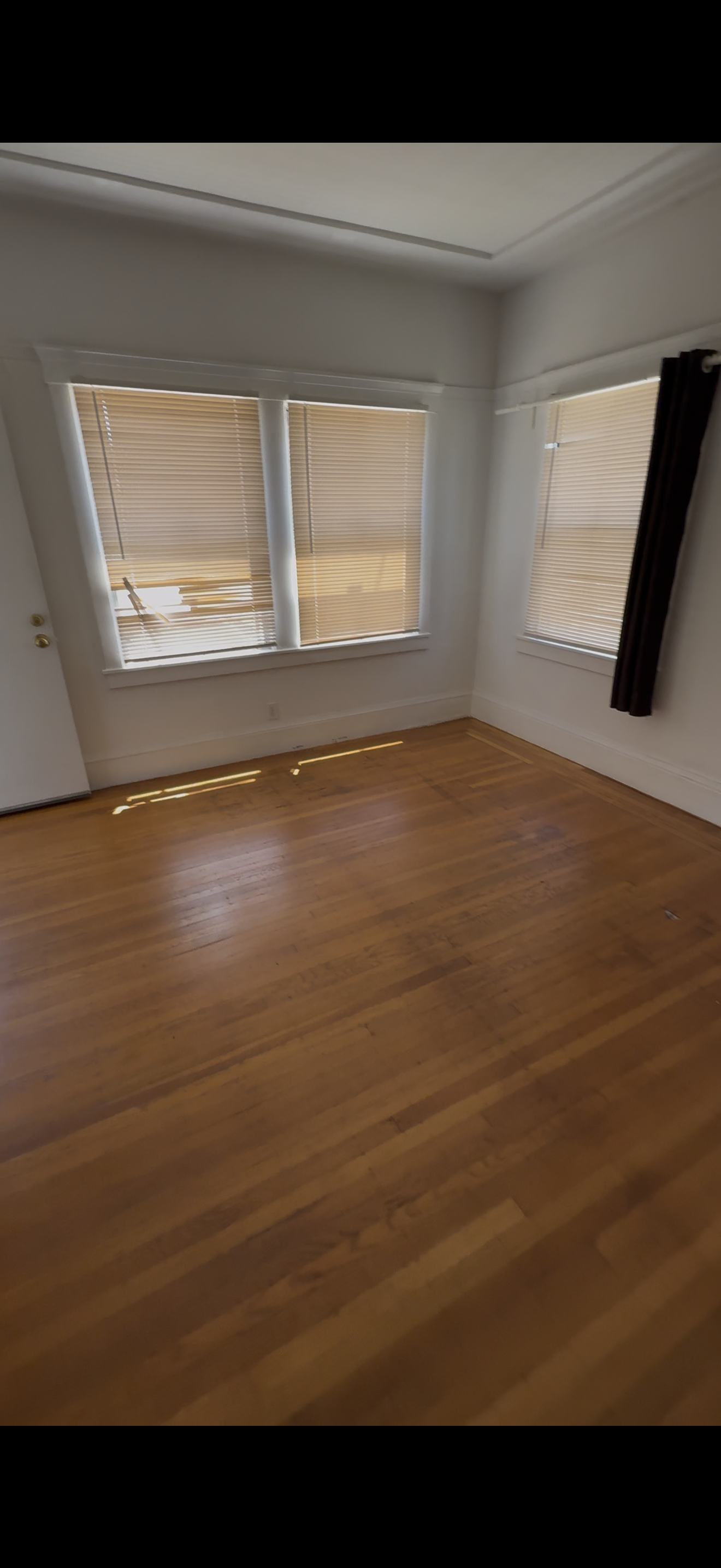 Empty room with hardwood floors, three large windows with blinds, and a black curtain on the right window.
