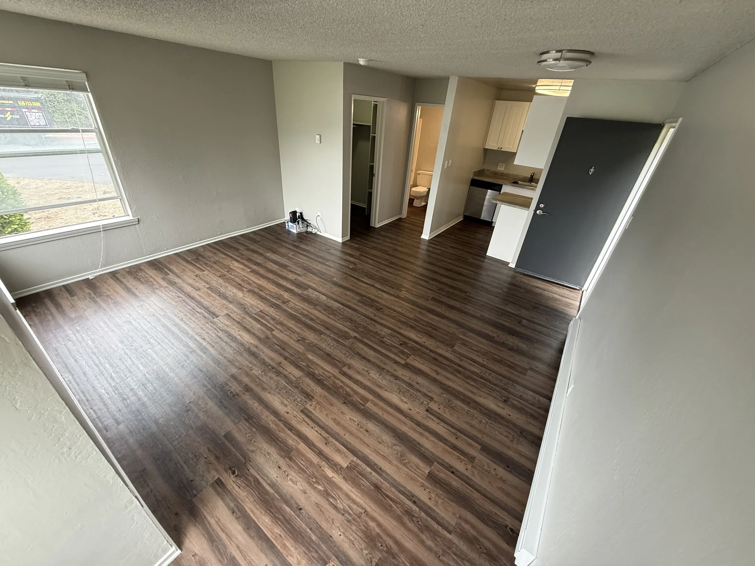 Empty room with wood-look flooring, a window to the left, and small kitchen area with beige cabinets and a stainless steel dishwasher to the right.