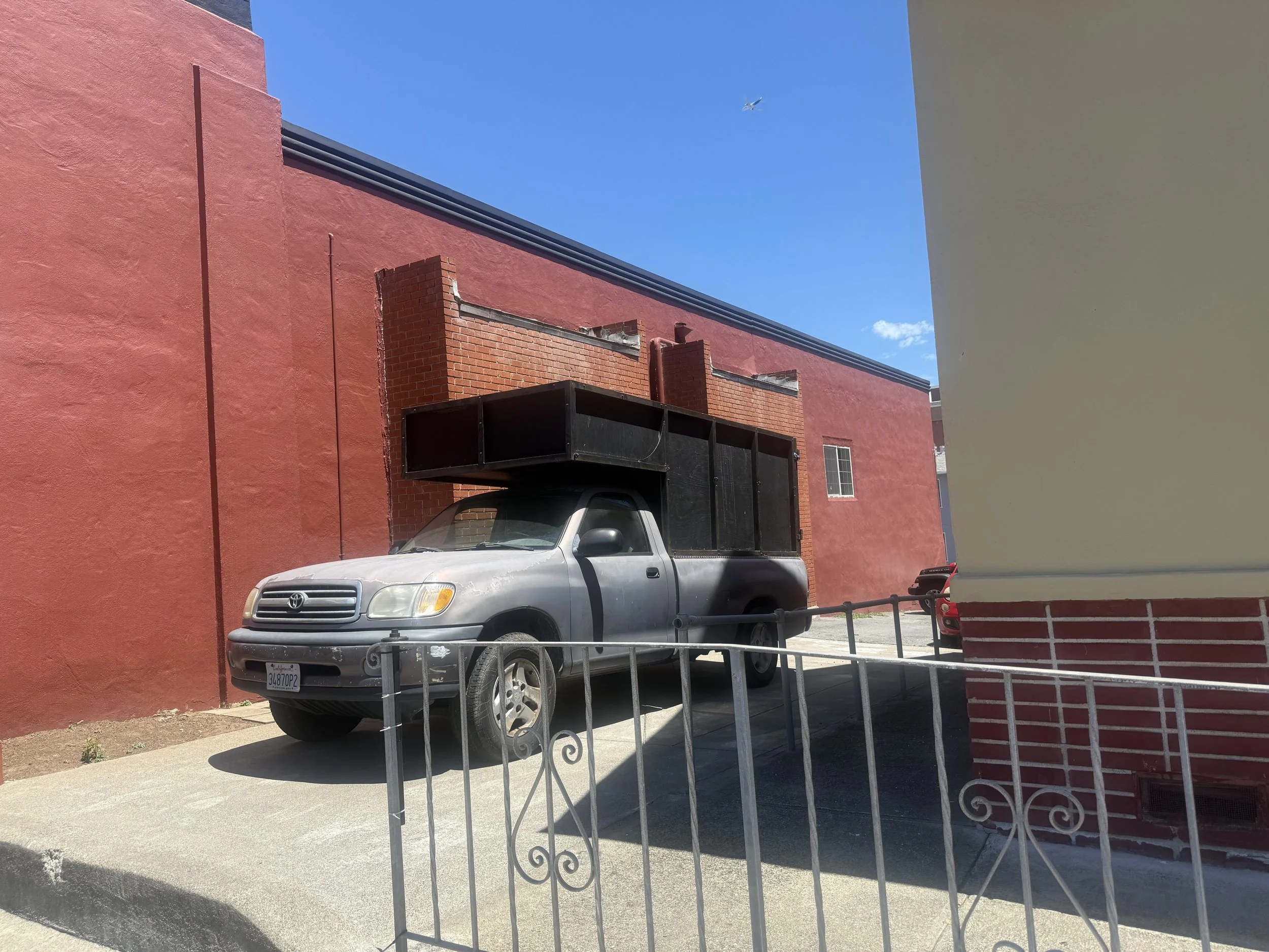 A silver Toyota pickup truck parked in front of a pink and red brick building, with blue sky and an airplane flying overhead. There is a small black structure attached to the truck and a metal railing in the foreground.