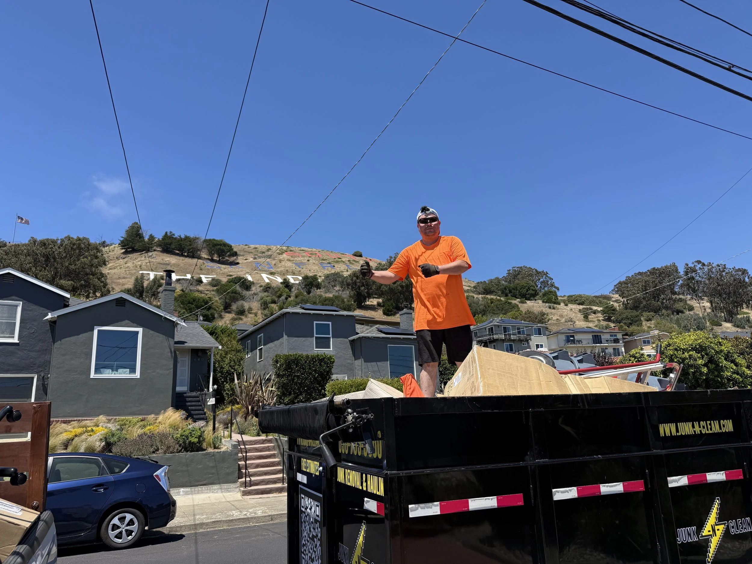 A man wearing an orange shirt, black shorts, gloves, and sunglasses standing on a truck bed filled with boxes and debris, with houses and a hillside in the background under a clear blue sky.