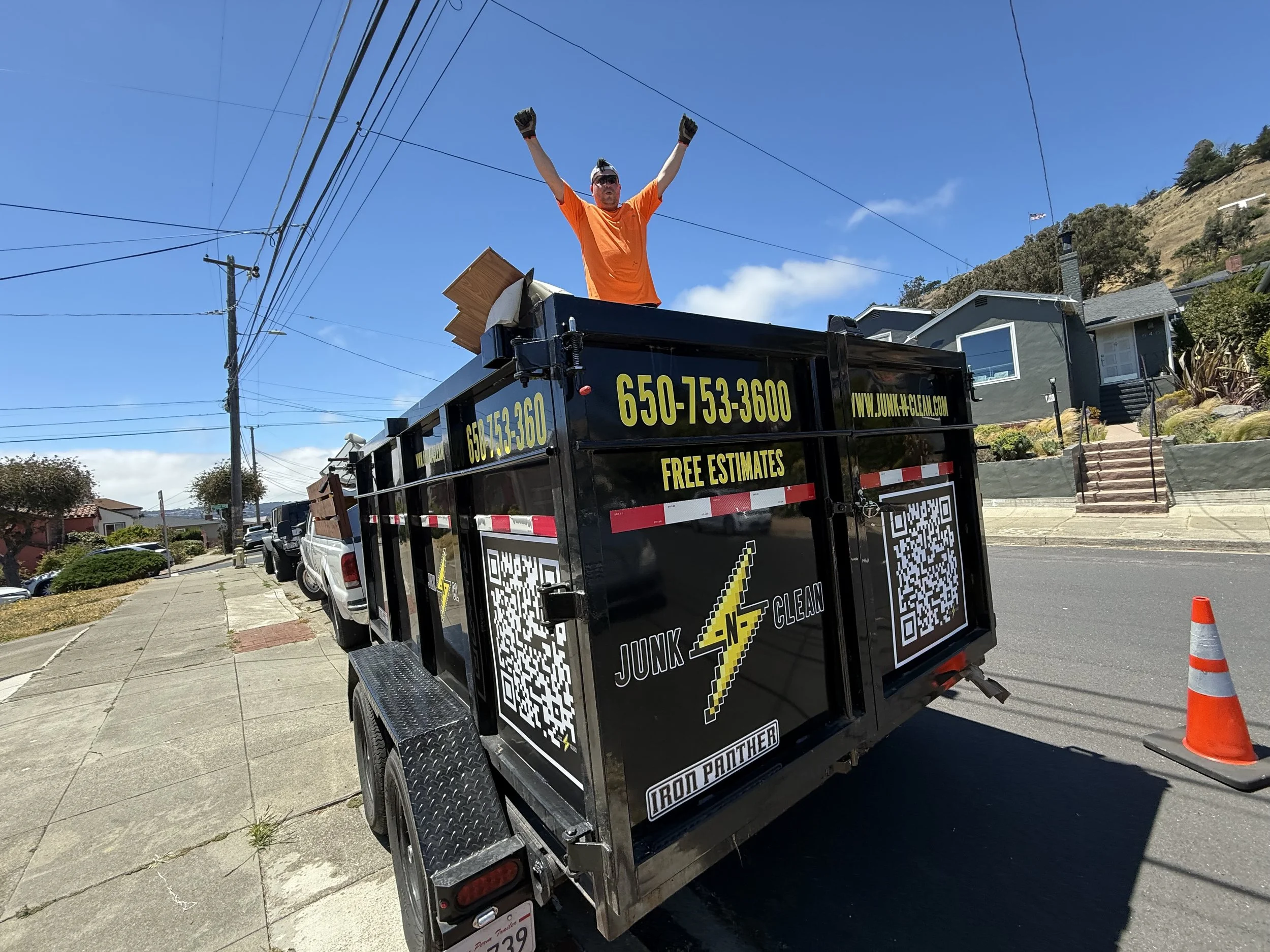 A man wearing an orange shirt and sunglasses standing in the back of a black junk removal truck with his arms raised in victory on a sunny day, with residential houses and power lines in the background.