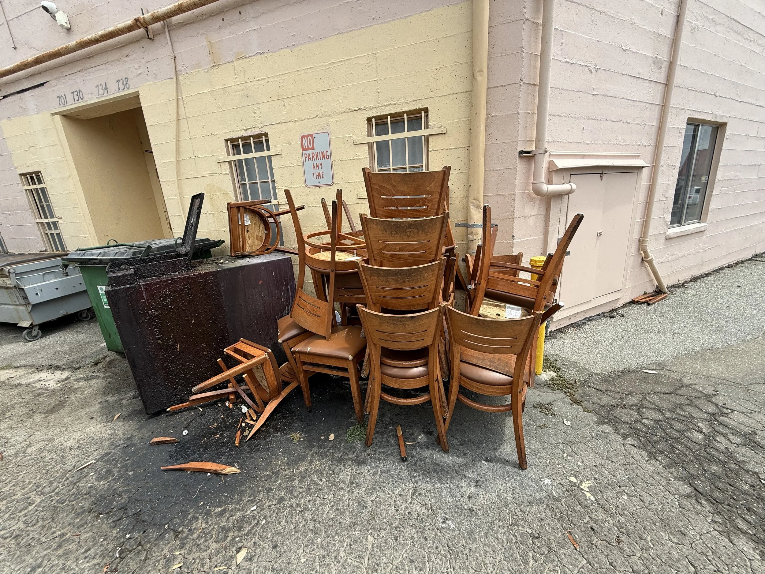 A pile of overturned wooden chairs leaning against a beige wall outside. The chairs are stacked with some legs and arms broken or missing. There are trash bins nearby, and a "No Parking Any Time" sign on the wall. The ground is a rough asphalt surfac