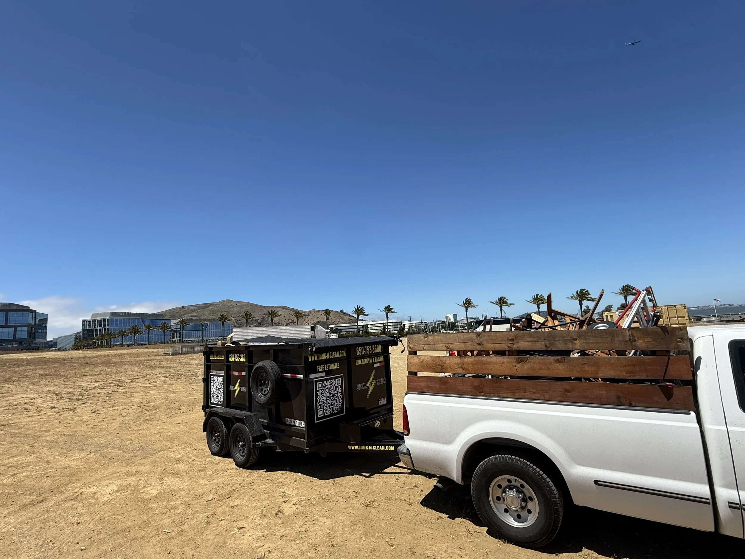 A pickup truck with a wooden bed loaded with miscellaneous items, and a black trailer with electronic and QR code labels in a dirt parking lot under blue sky, with modern buildings and palm trees in the background.