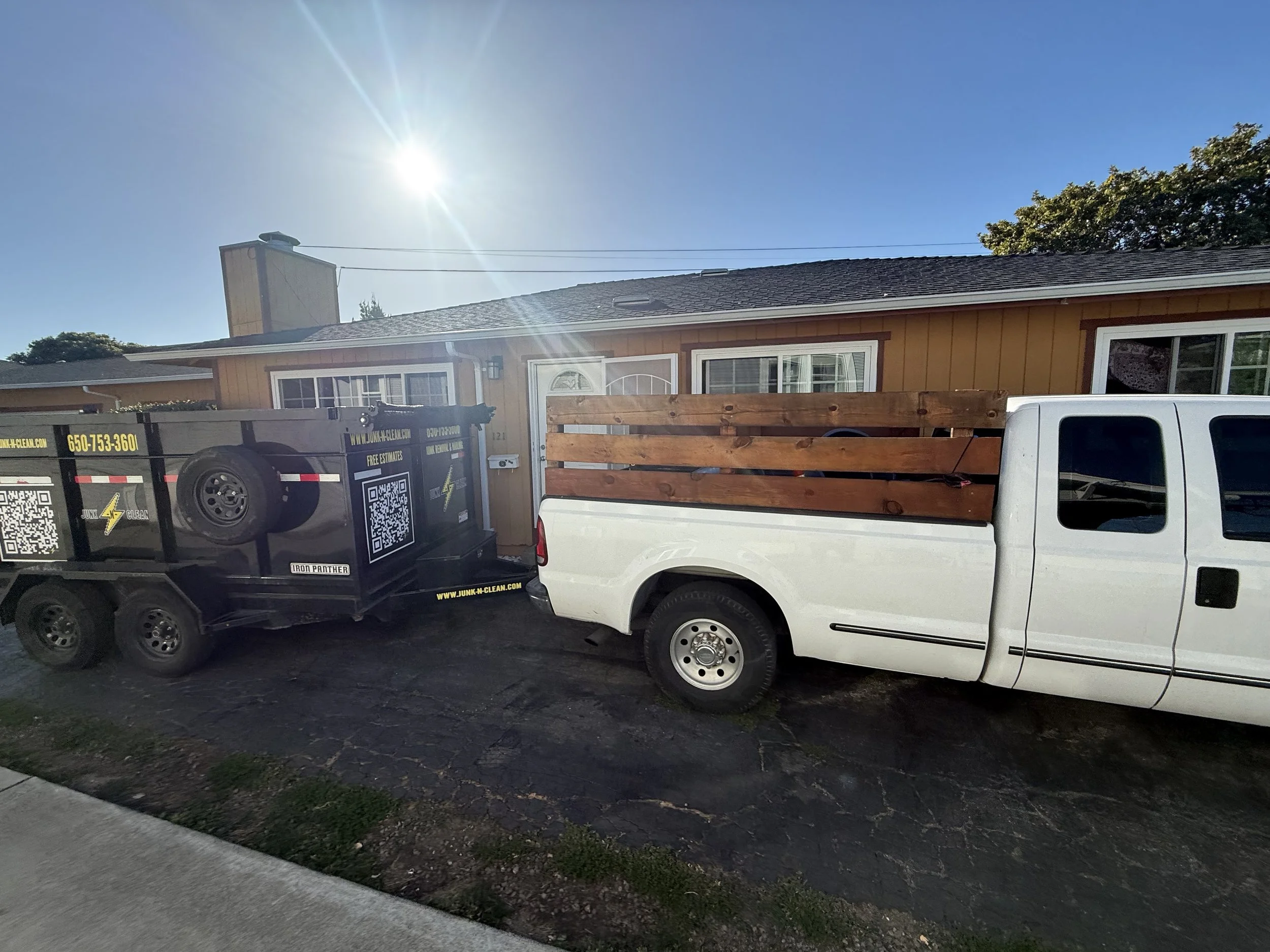 A white pickup truck parked in front of a house with a wooden fence and a large trash bin nearby, under a clear blue sky with the sun shining brightly.