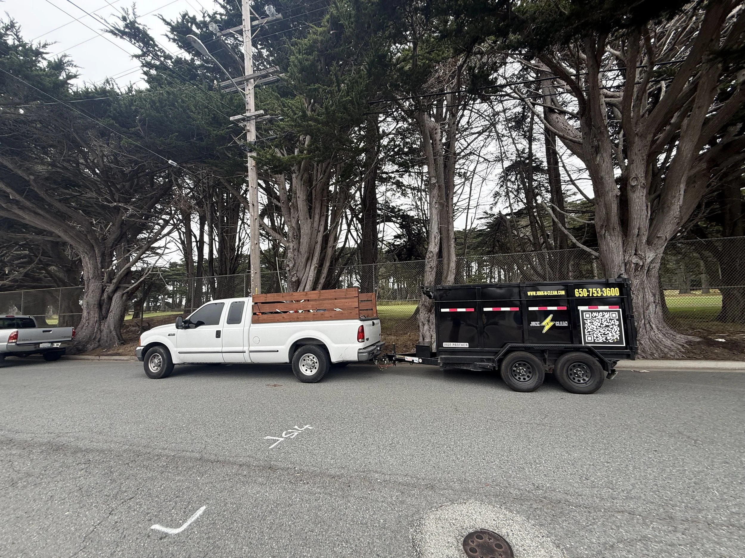 Parked vehicles, trees, power lines, and a waste disposal trailer in a parking lot.