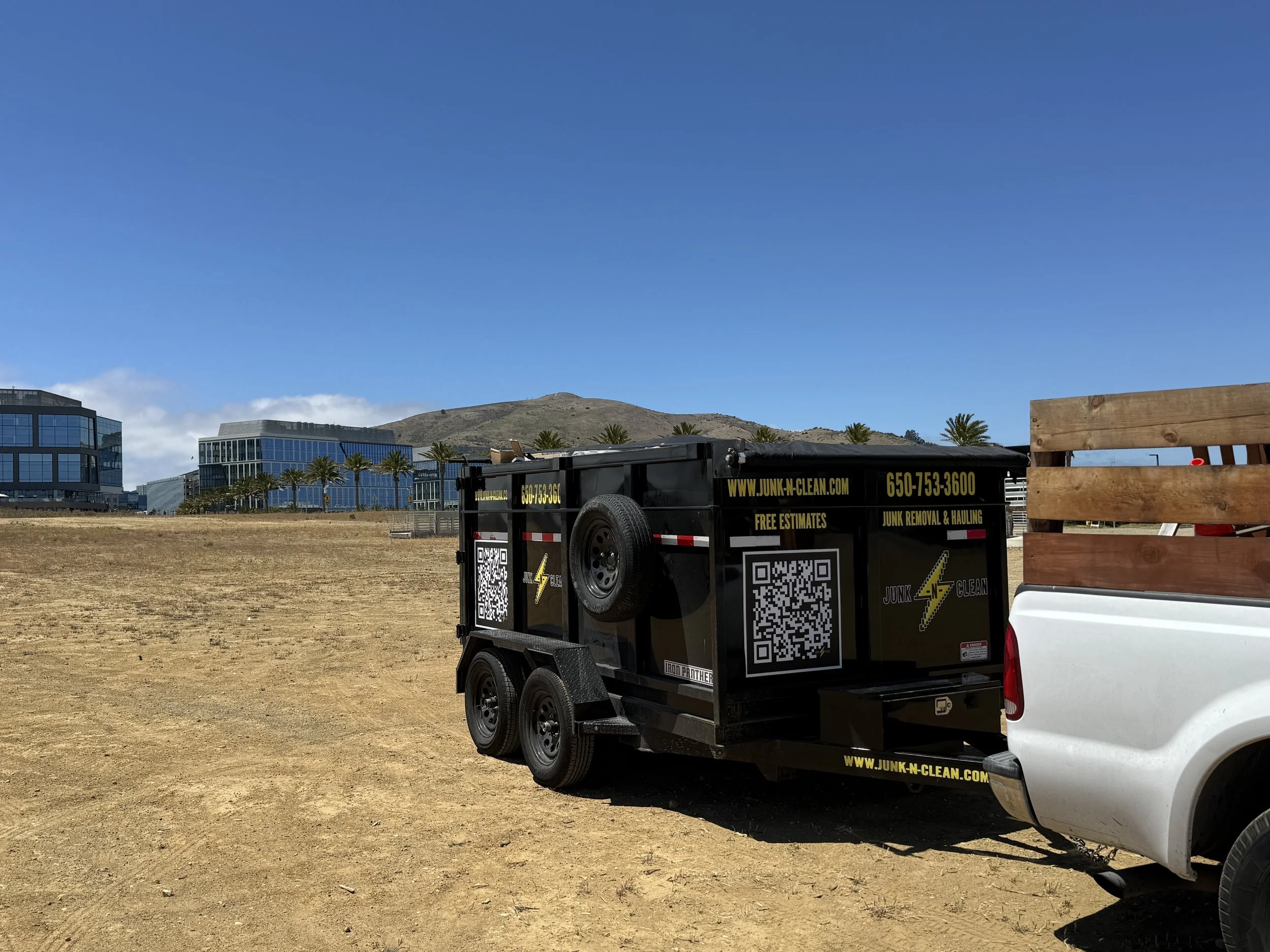 A black trailer attached to a white pickup truck parked on a dirt field, with a background of modern glass buildings, palm trees, and a mountain under a partly cloudy sky.