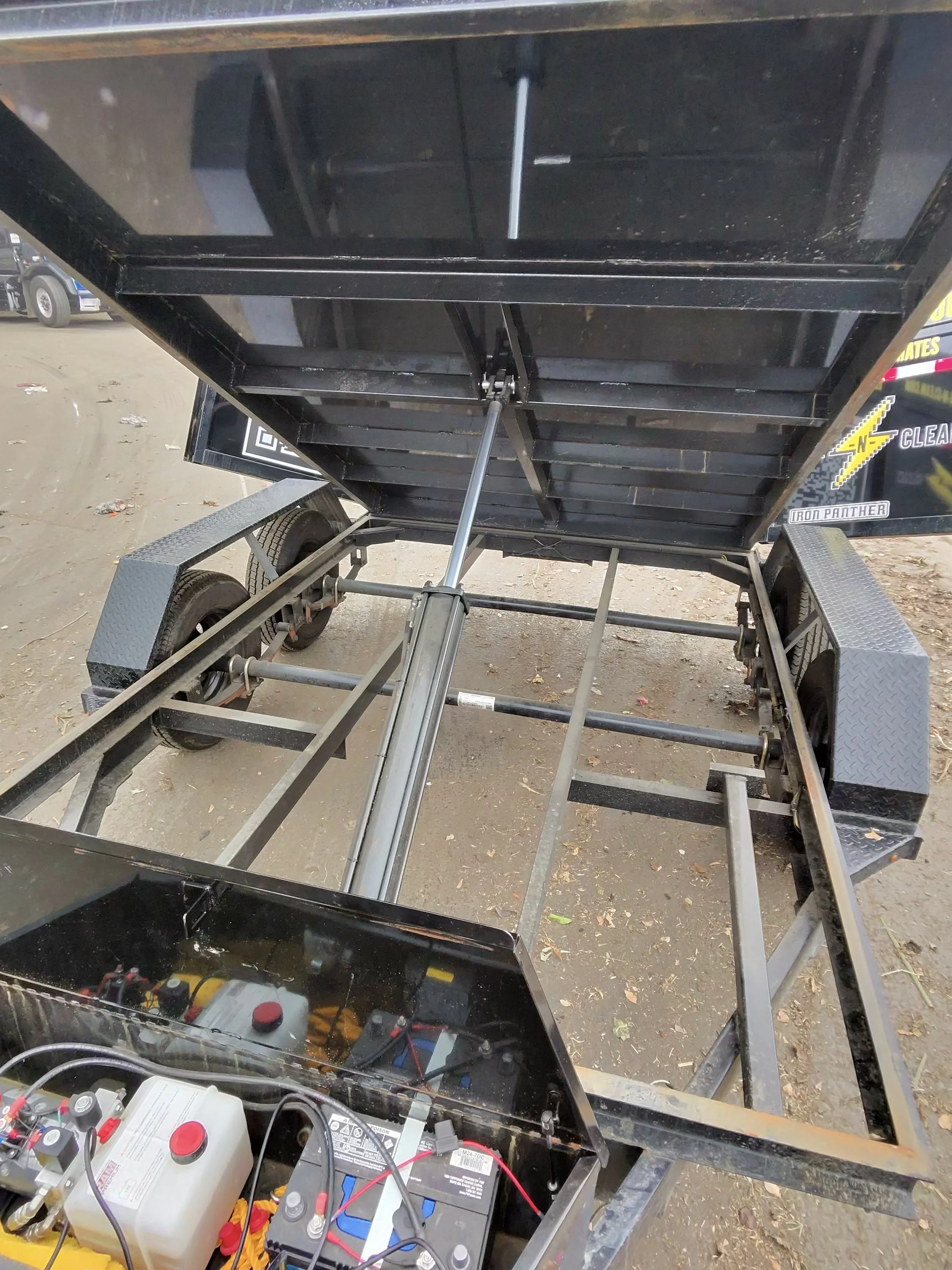 The photo shows a black metal frame structure with an open top, connected to a handle and wheel assembly. Inside, there are batteries, wiring, and electronic components. The background shows a parking lot with some vehicles.