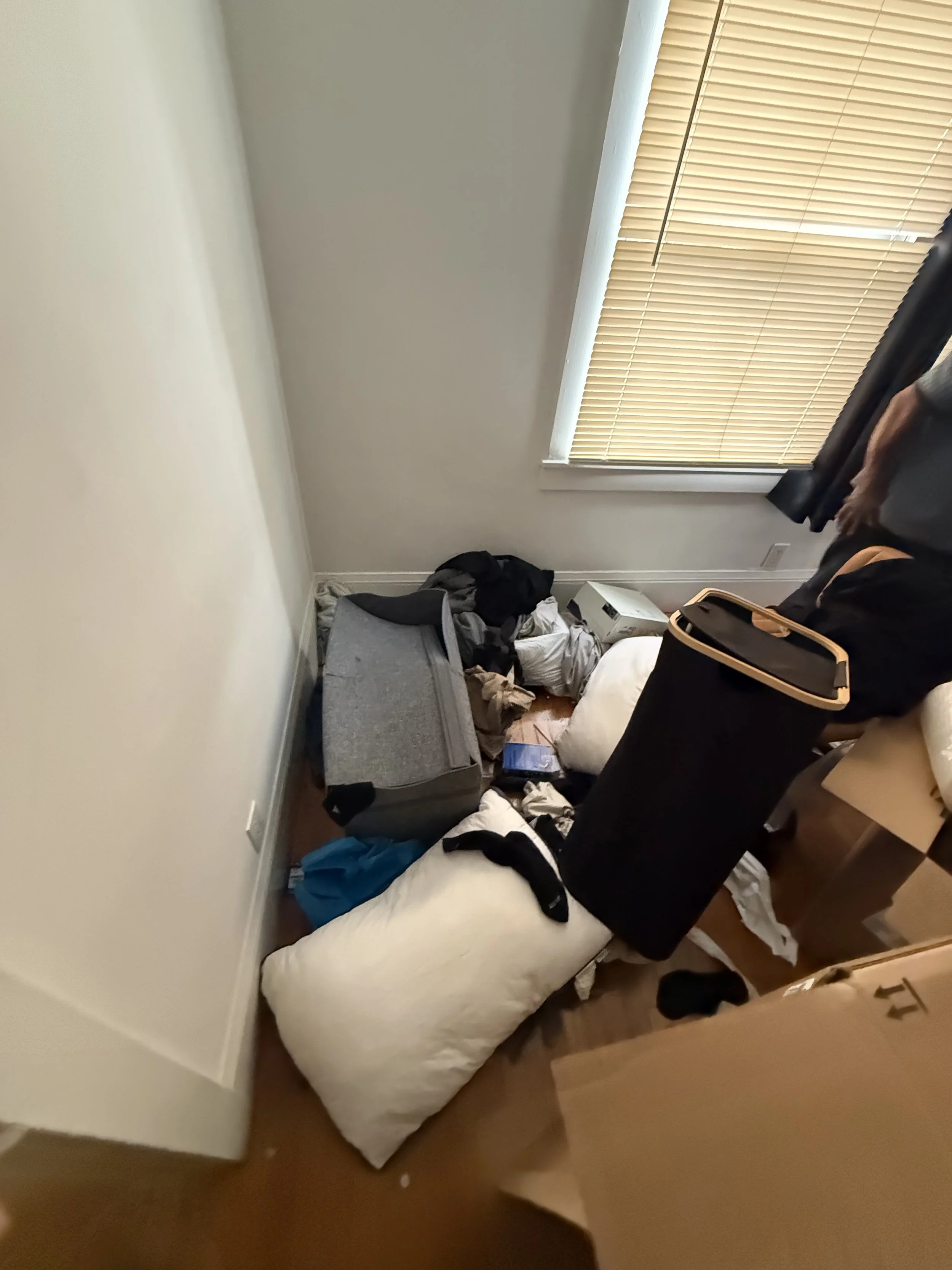 A cluttered bedroom with a mattress on the floor, filled with scattered clothing, bags, and miscellaneous items. There is a window with closed blinds, and a small wooden table partially visible. The room has white walls and hardwood flooring.