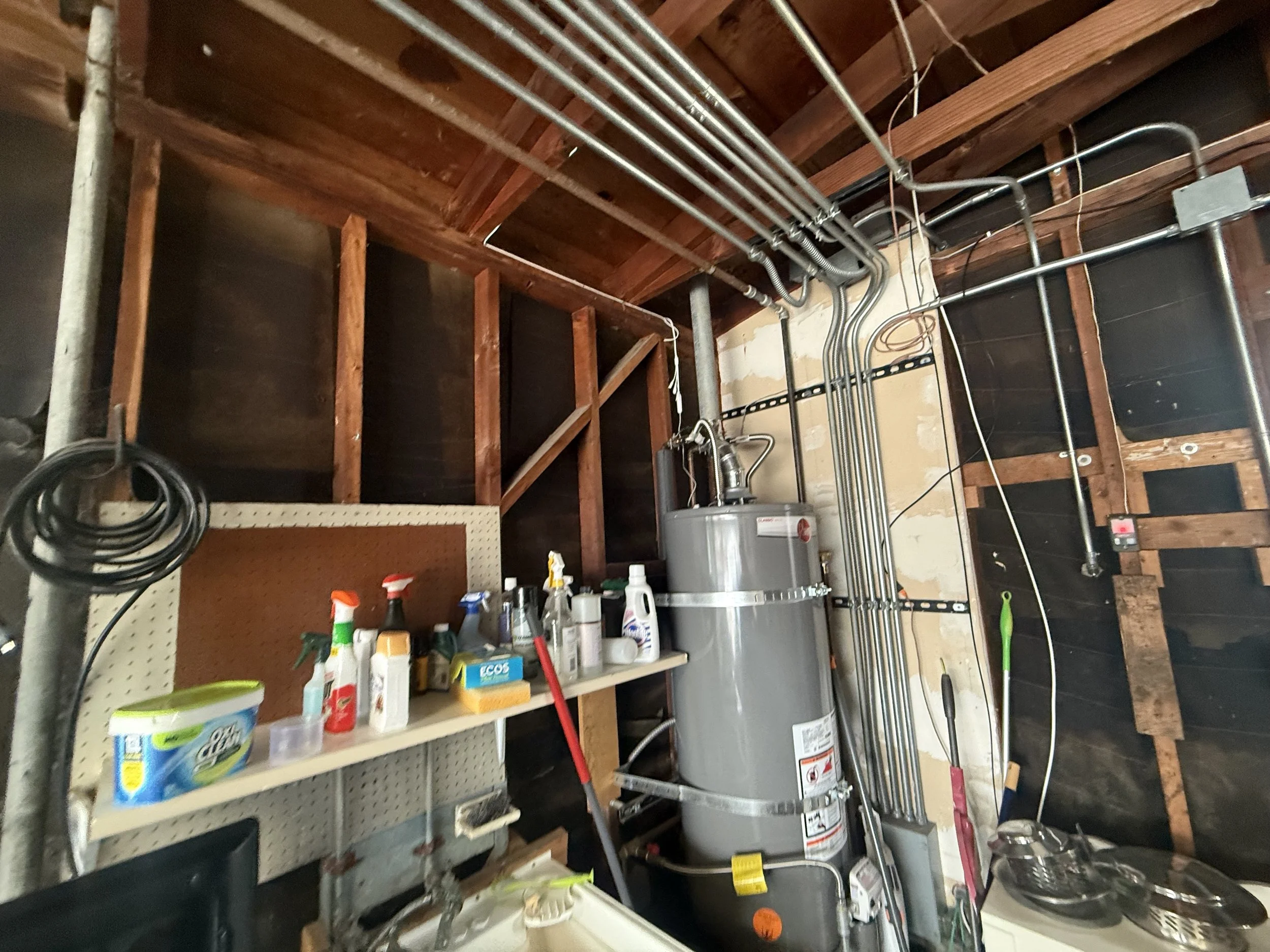 Garage or utility room with exposed wooden beams and electrical wiring. A water heater is visible along with cleaning supplies and tools on shelves. Hoses and a mop lay against the wall.