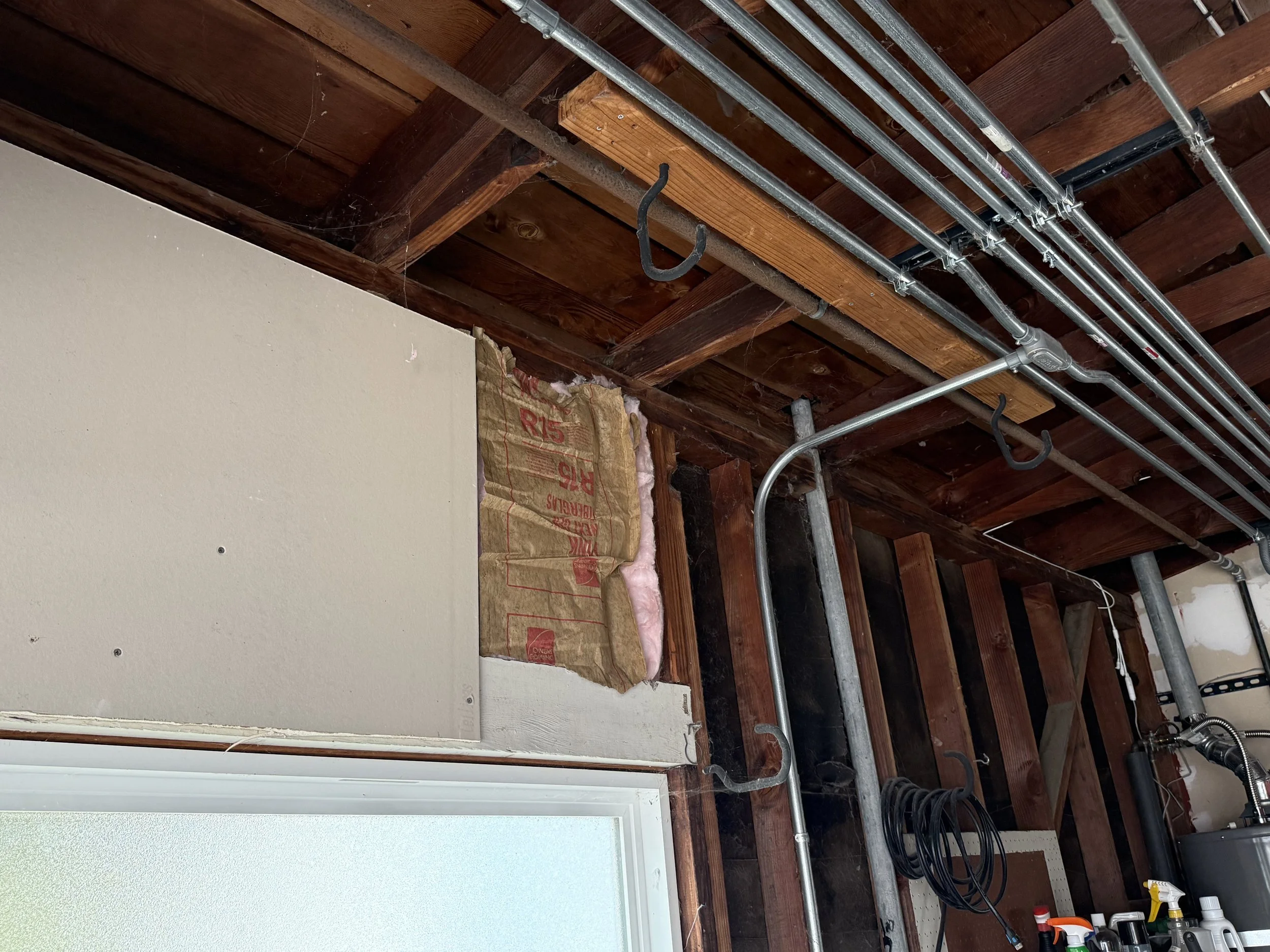 View of a ceiling with exposed wooden beams, metal pipes, and electrical wiring in a construction or renovation site. Some insulation and drywall are visible.