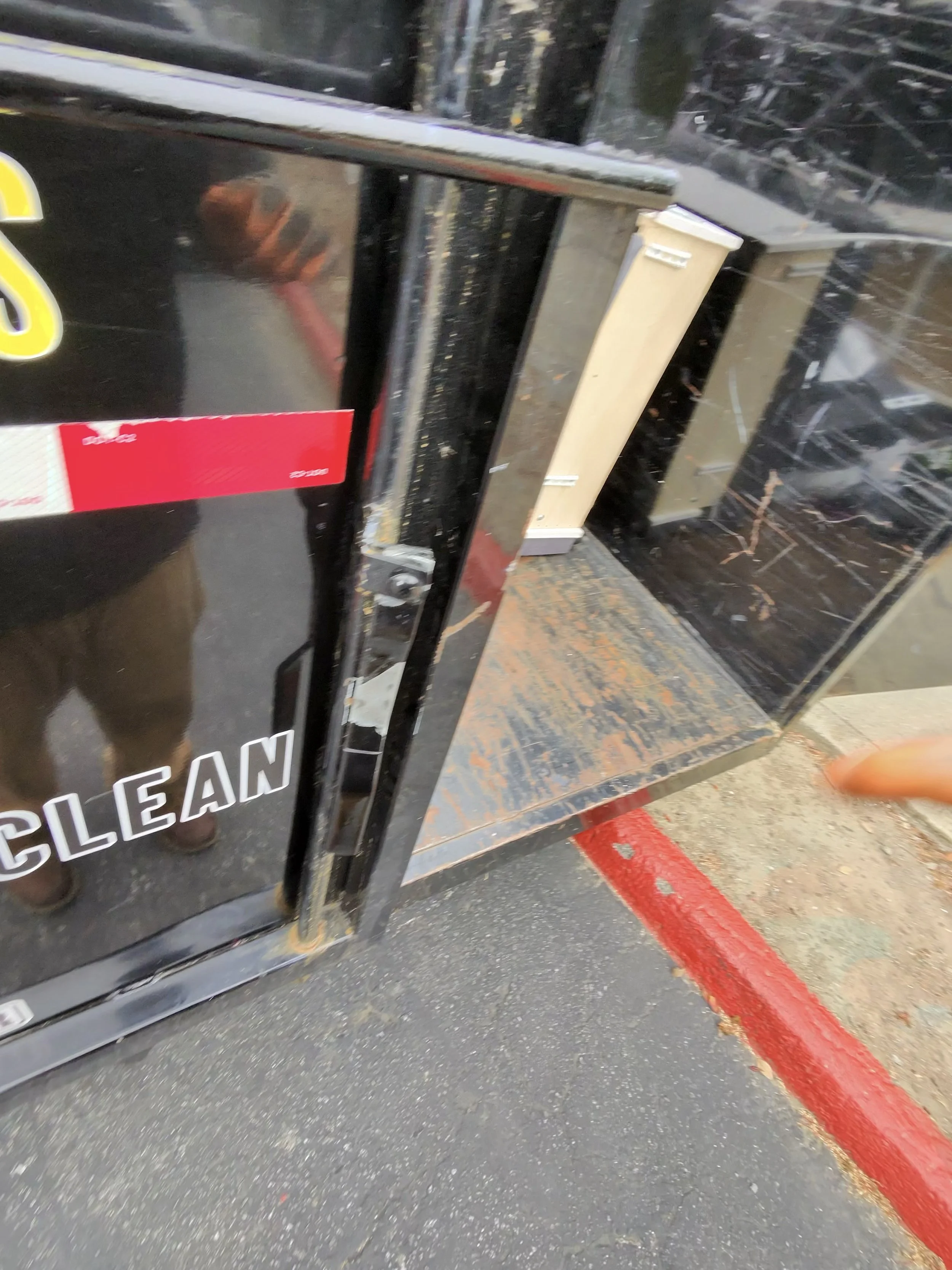 A black glass door with white and red label tape, showing a reflection of a person taking the photo, and a red painted curb outside.