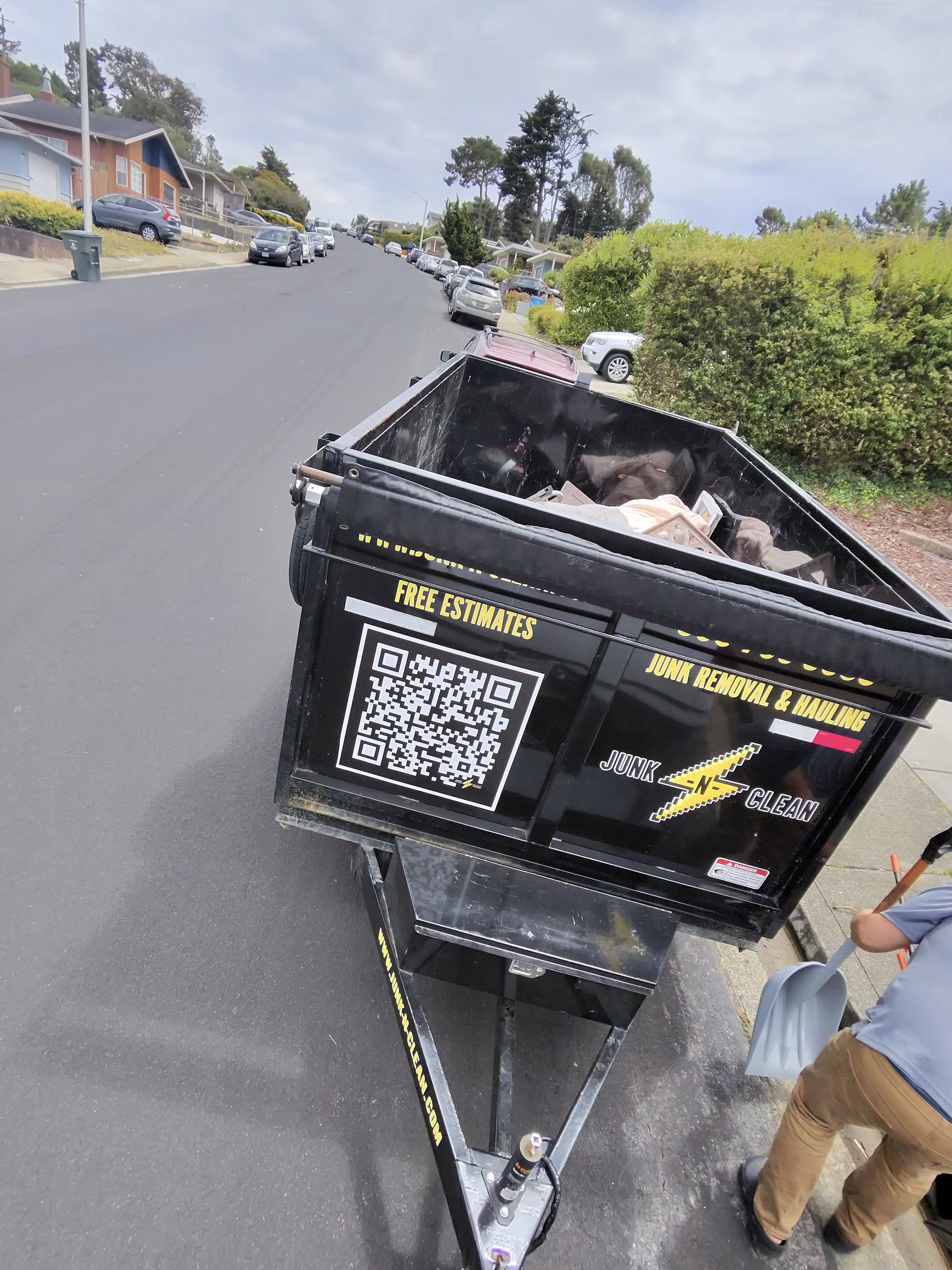 View of a street with parked cars, houses, and trees in a neighborhood. In the foreground, part of a black junk removal and hauling cart with a QR code and promotional text is visible, along with a person wearing brown pants and a gray shirt.