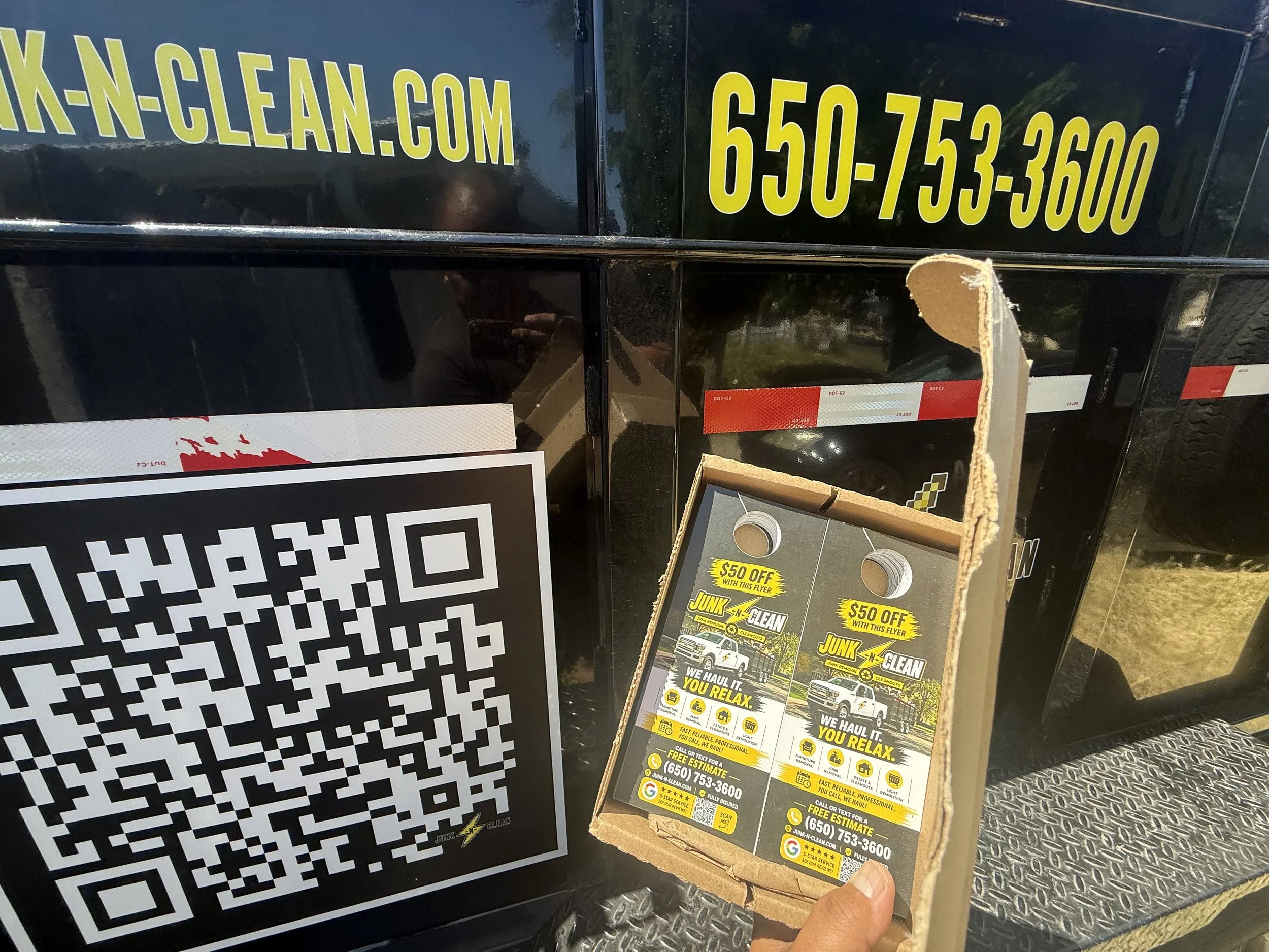 Close-up of a phone number, website URL, and promotional flyer for a junk removal service, held by a person hand. Part of a QR code is visible in the background.