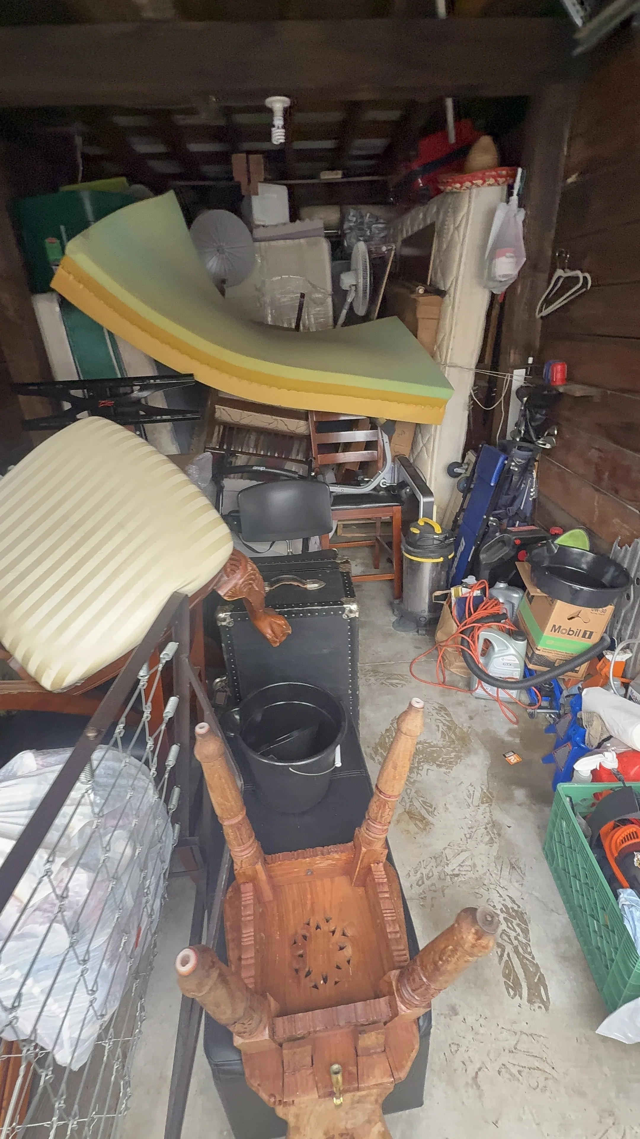 A cluttered storage space with various furniture items, tools, and household objects haphazardly arranged.