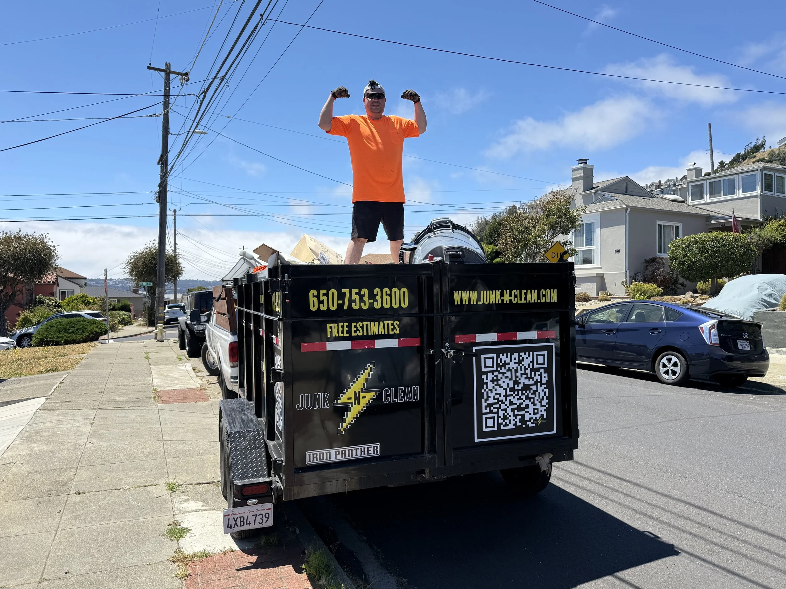 A man in an orange shirt and black shorts standing on a truck bed, flexing his arms. The truck has a sign with contact details for junk removal and a QR code. The background features residential houses, power lines, and a blue sky with some clouds.