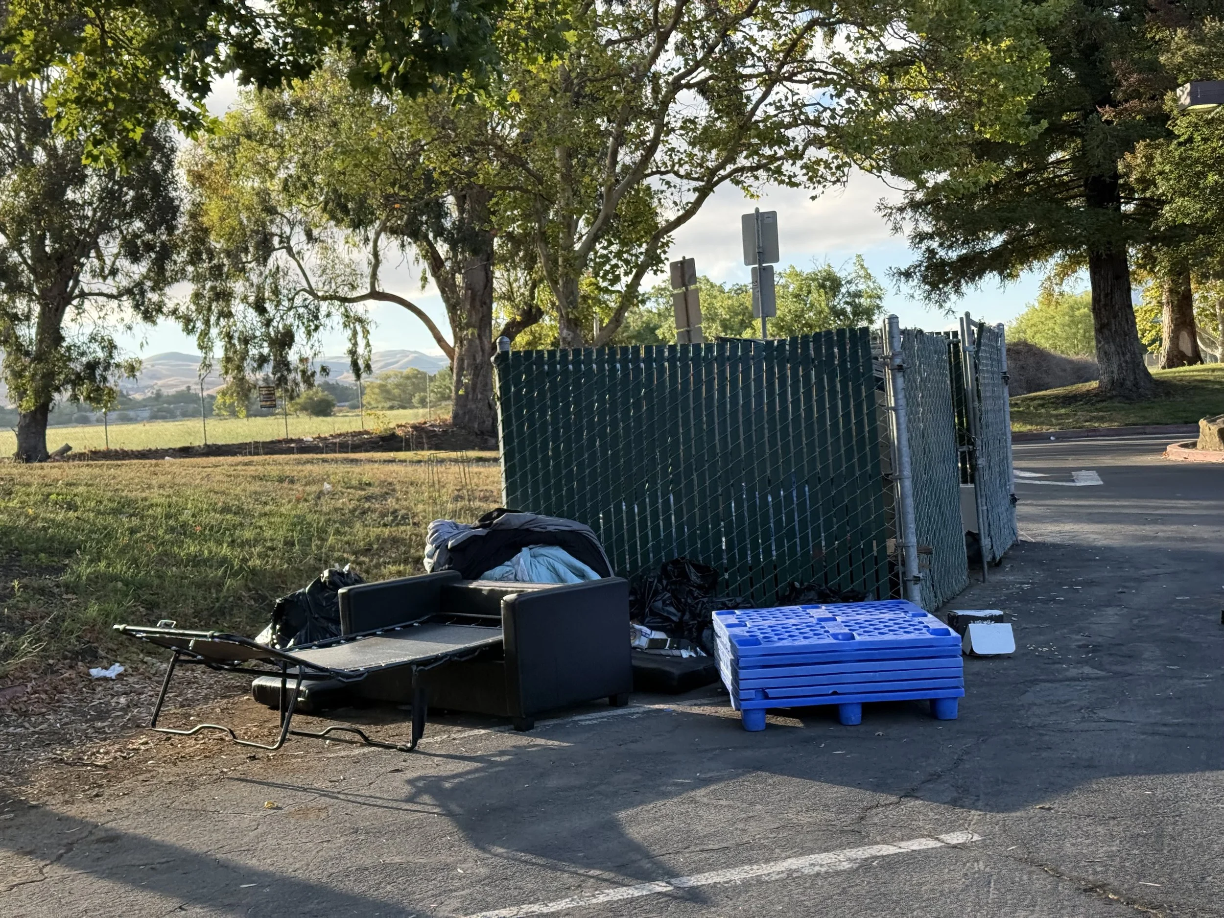A parking lot with a green chain-link fence, trees, and a scenic background of hills. There is discarded clothing and equipment on the ground, including a black folding chair, a blue plastic pallet, and some bags.