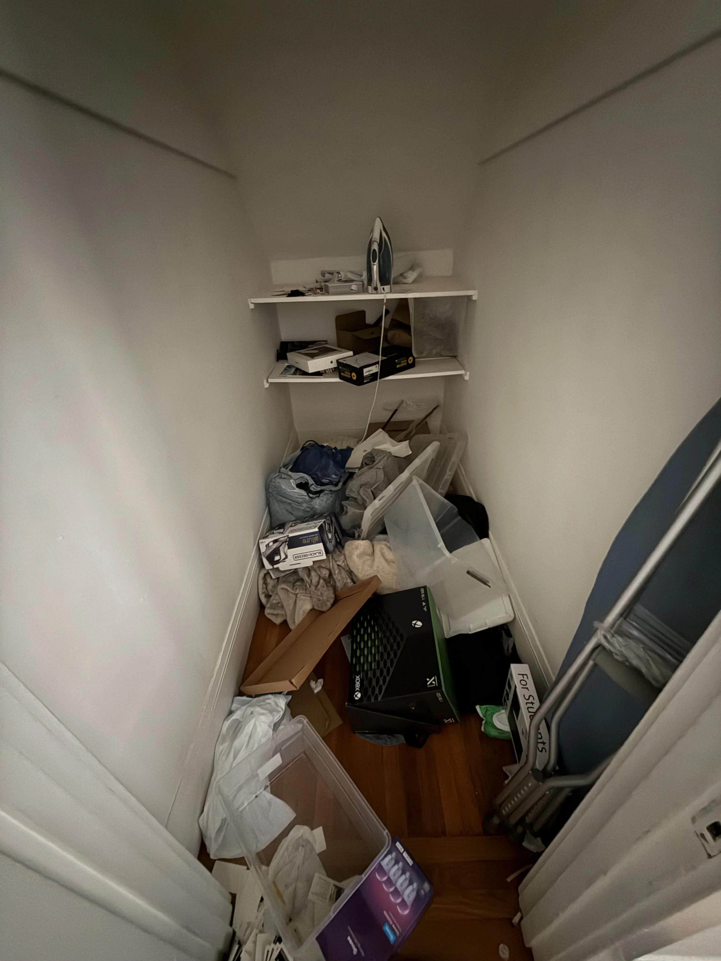 Cluttered closet space with scattered papers, boxes, an iron, a folding chair, and various household items.