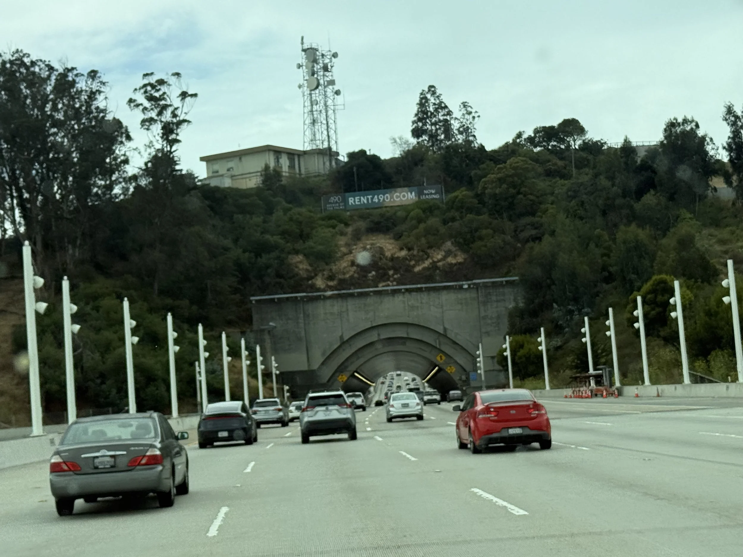 A highway with cars driving into a tunnel through a hillside covered with trees. There is a sign for rent490.com visible on the hillside above the tunnel.