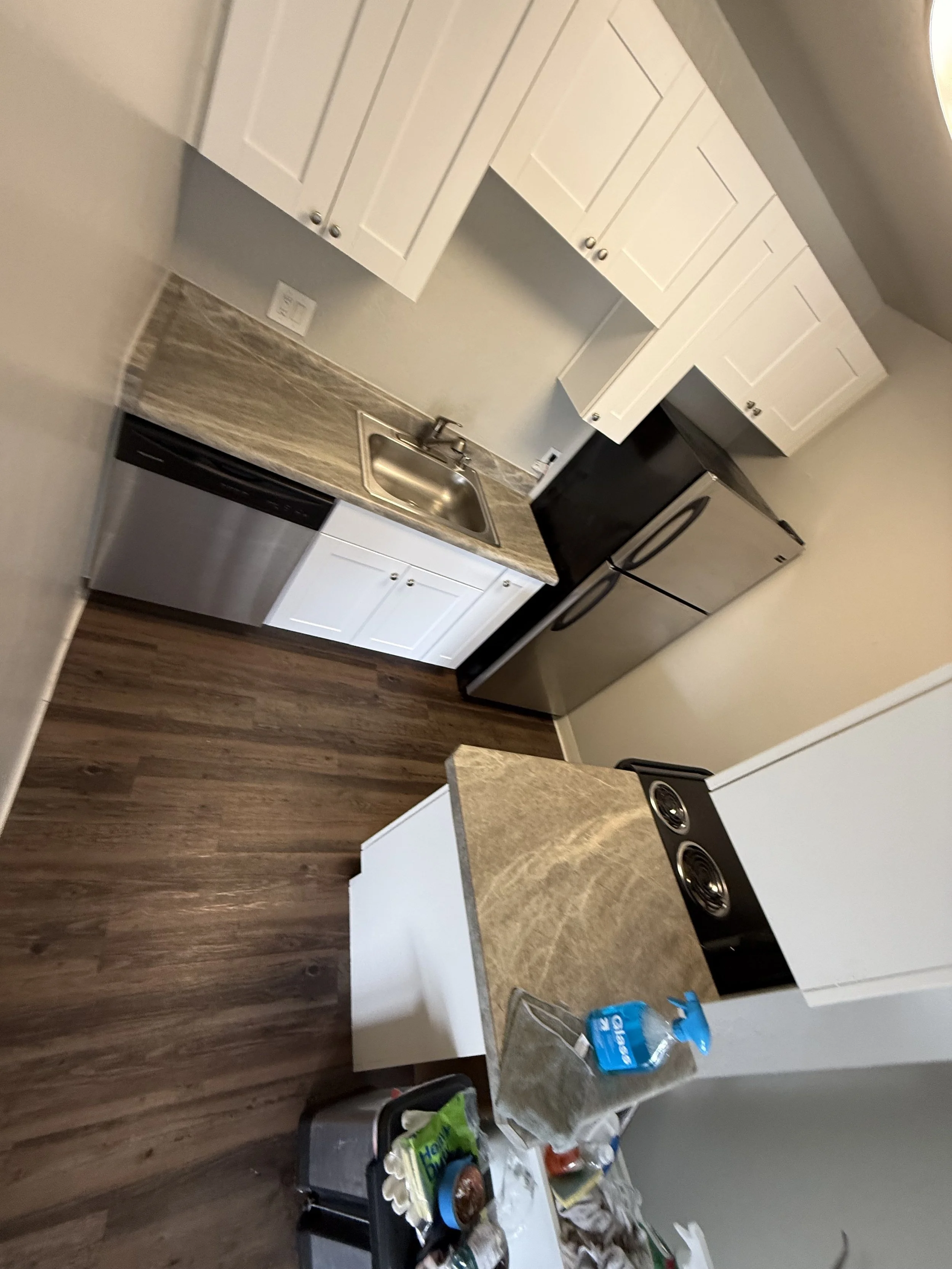 A small kitchen with white cabinets, a stainless steel sink, a black and stainless steel refrigerator, and a black stove. There is a countertop with various items including a box of chicken broth, a bag of chips, a plastic container, and some paper t