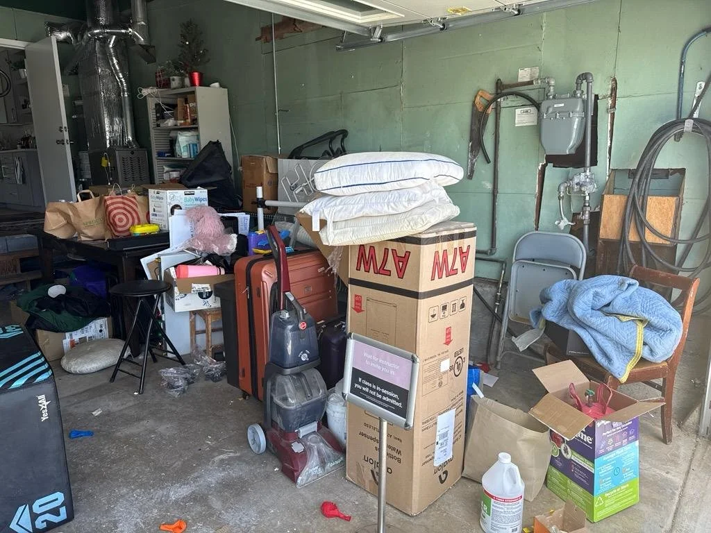 Cluttered garage with various household items including boxes, luggage, pillows, a vacuum cleaner, chairs, and miscellaneous objects.