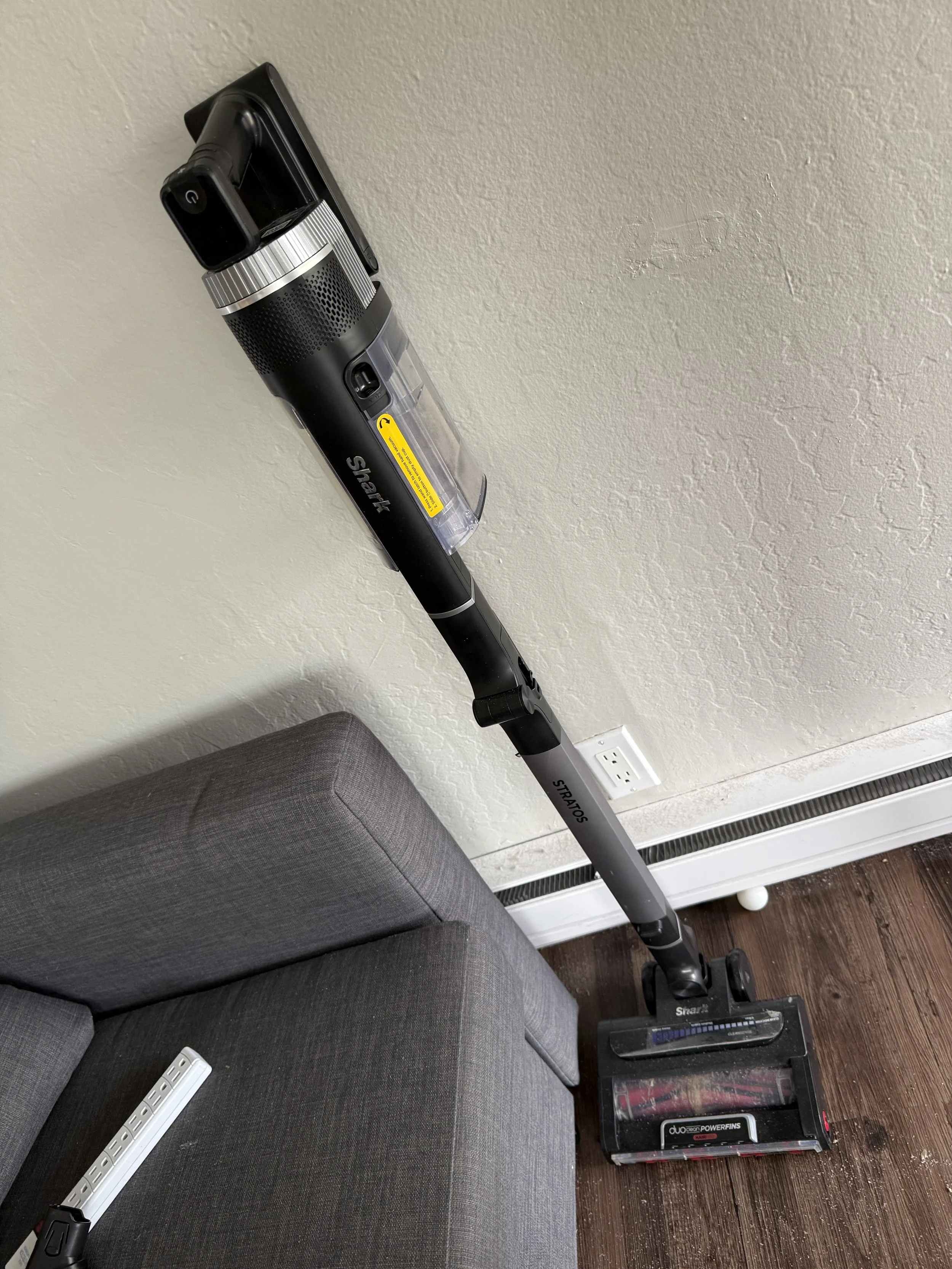 A cordless vacuum cleaner leaning against a sofa near a baseboard heater.