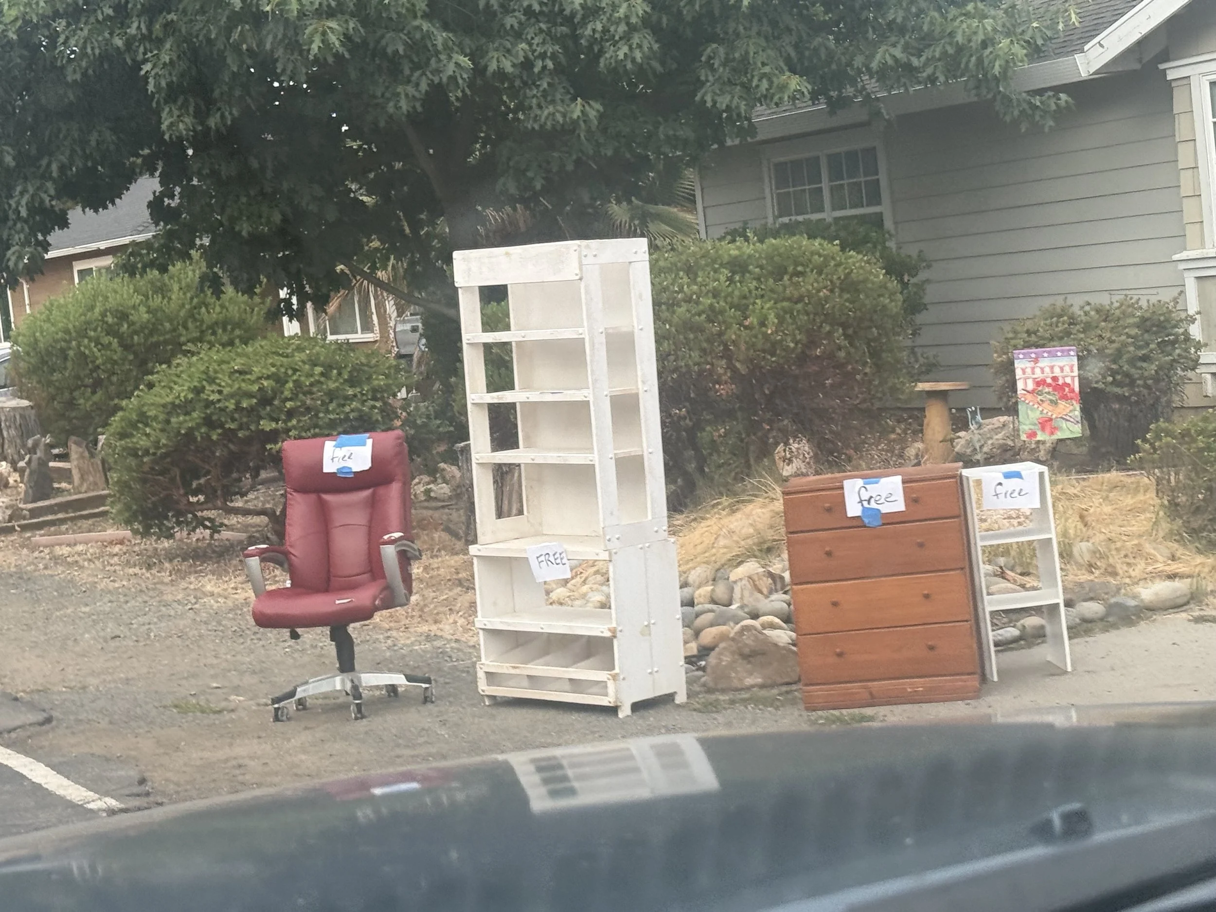 Various free items for donation such as a chair, bookshelf, and small furniture, displayed outside a house with plants and rock landscaping.