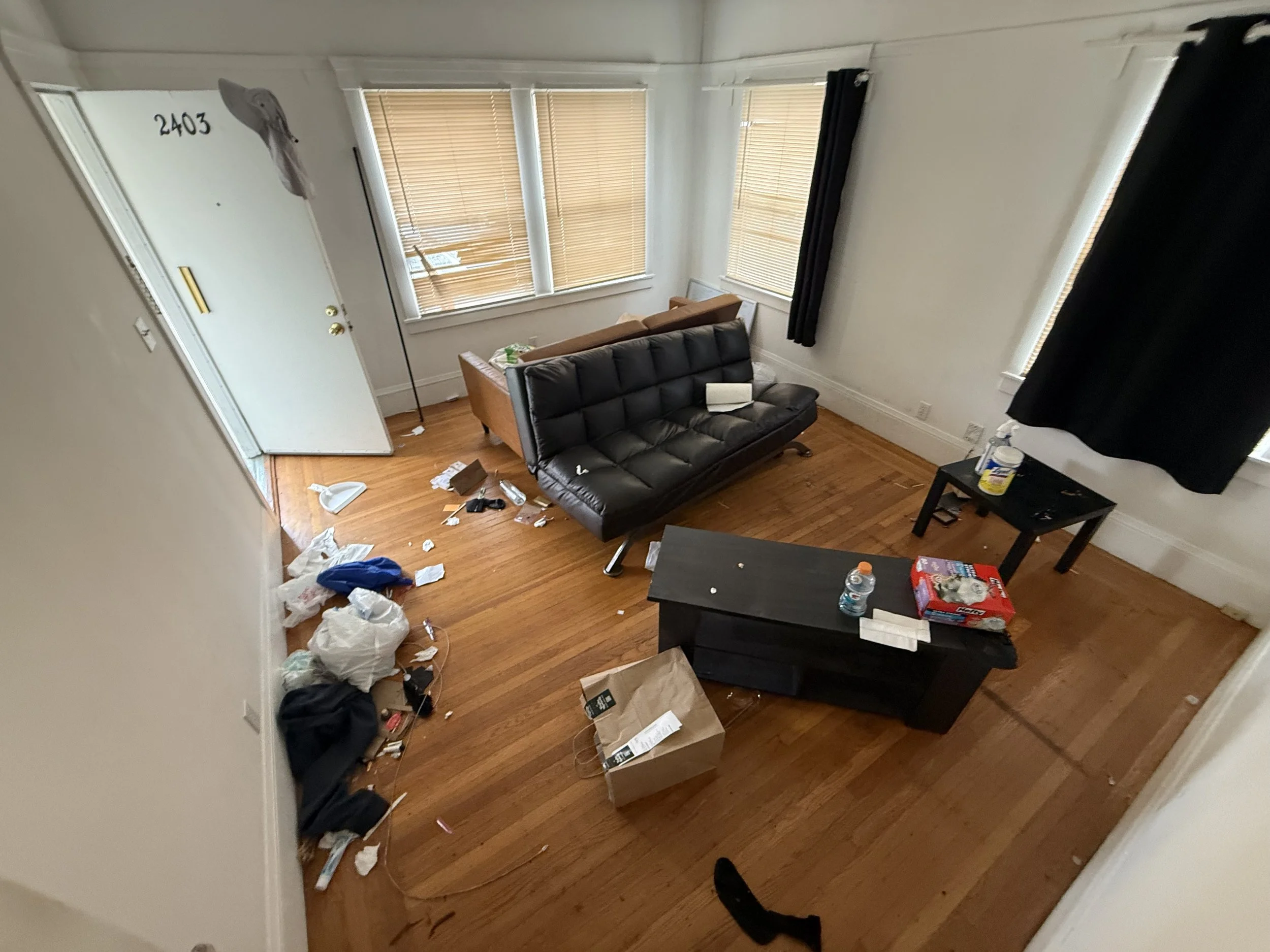 A messy living room with scattered trash, cardboard boxes, and paper. Sofa and windows with blinds are visible.