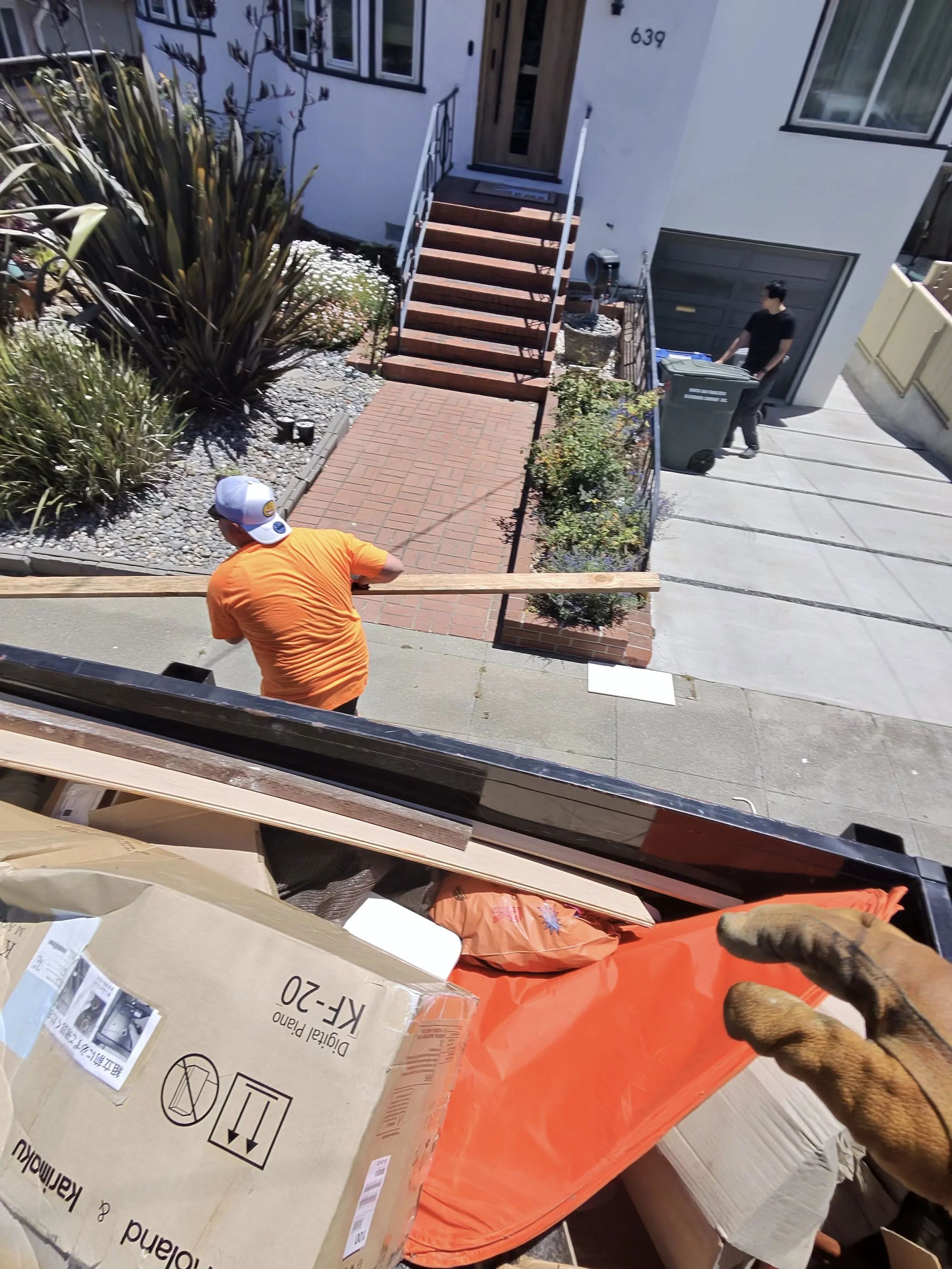 A man in an orange shirt and white cap working with cardboard boxes inside a house, at an upper level looking down, with another man in black clothes outside near a green trash bin, and a dog with a brown coat visible in the bottom right corner.