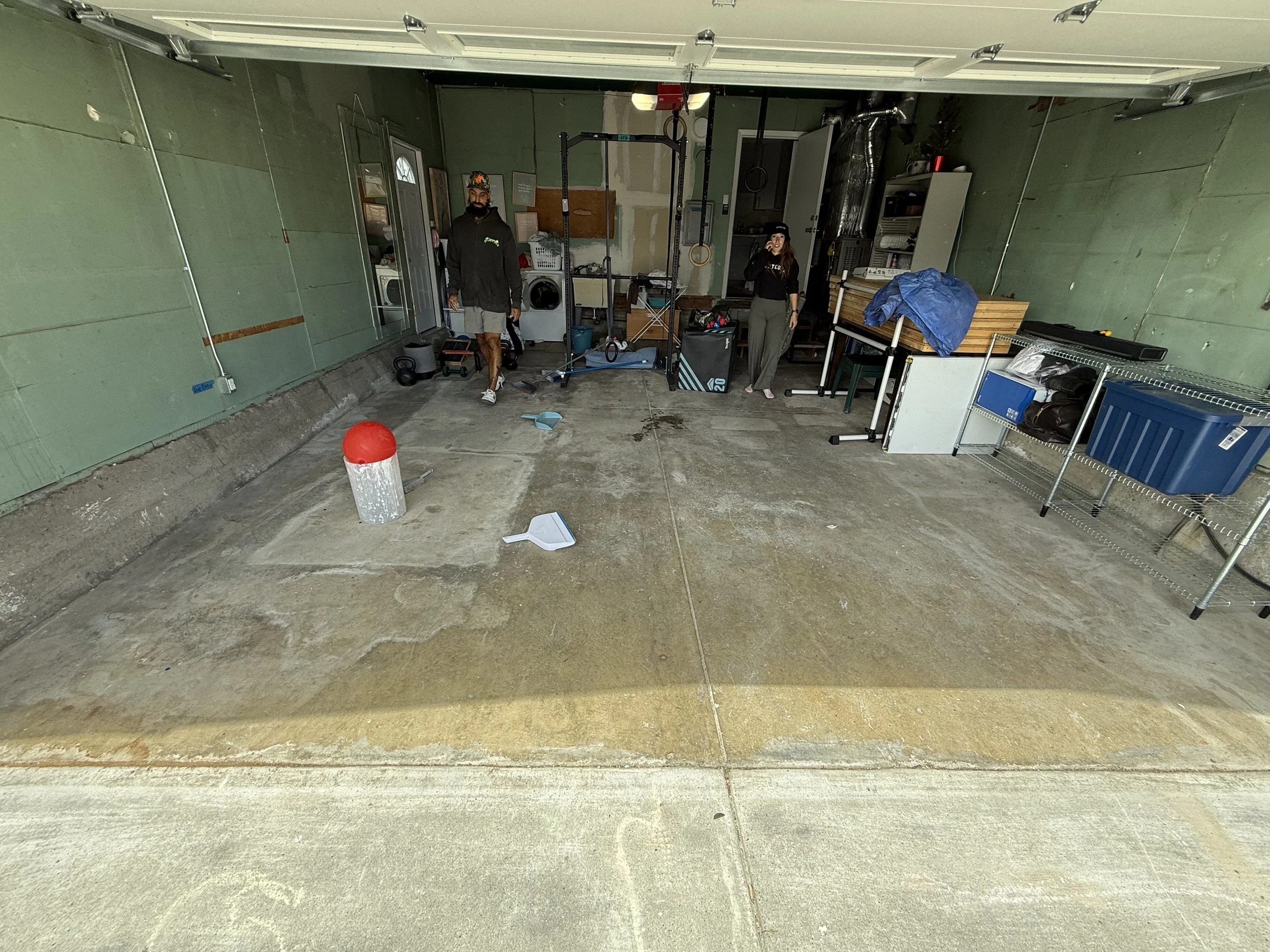 A garage with two people standing inside, with various workout equipment, storage shelves, and a washing machine visible. The floor is concrete, with some dirt and a dustpan and broom in the foreground.
