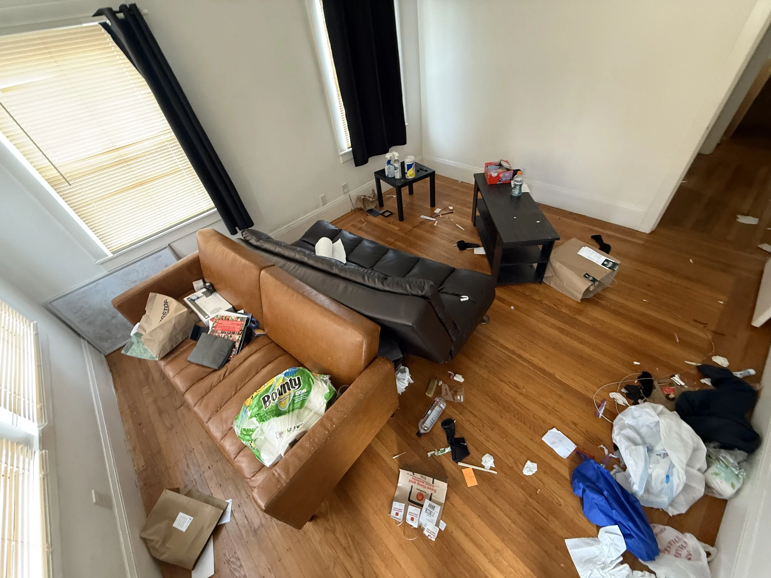 Disorganized living room with scattered trash including paper, boxes, and plastic bags. Couch with some items on it, small tables holding various objects, and windows with blinds and black curtains.